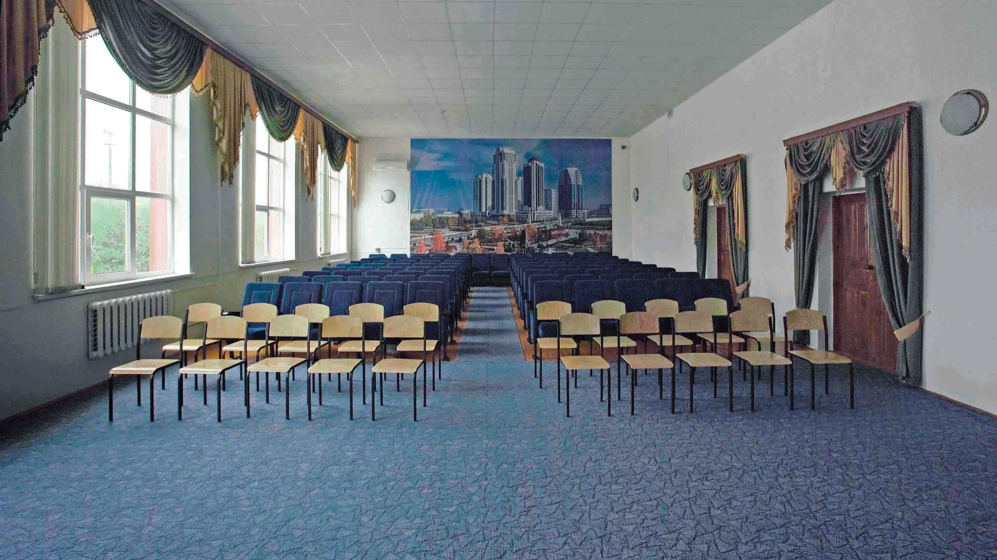 An image of a blue carpeted room filled with rows of chairs. A large photograph of a cityscape fills the back wall. Windows line the left wall, two doors are on the right. Both have ornate drapery