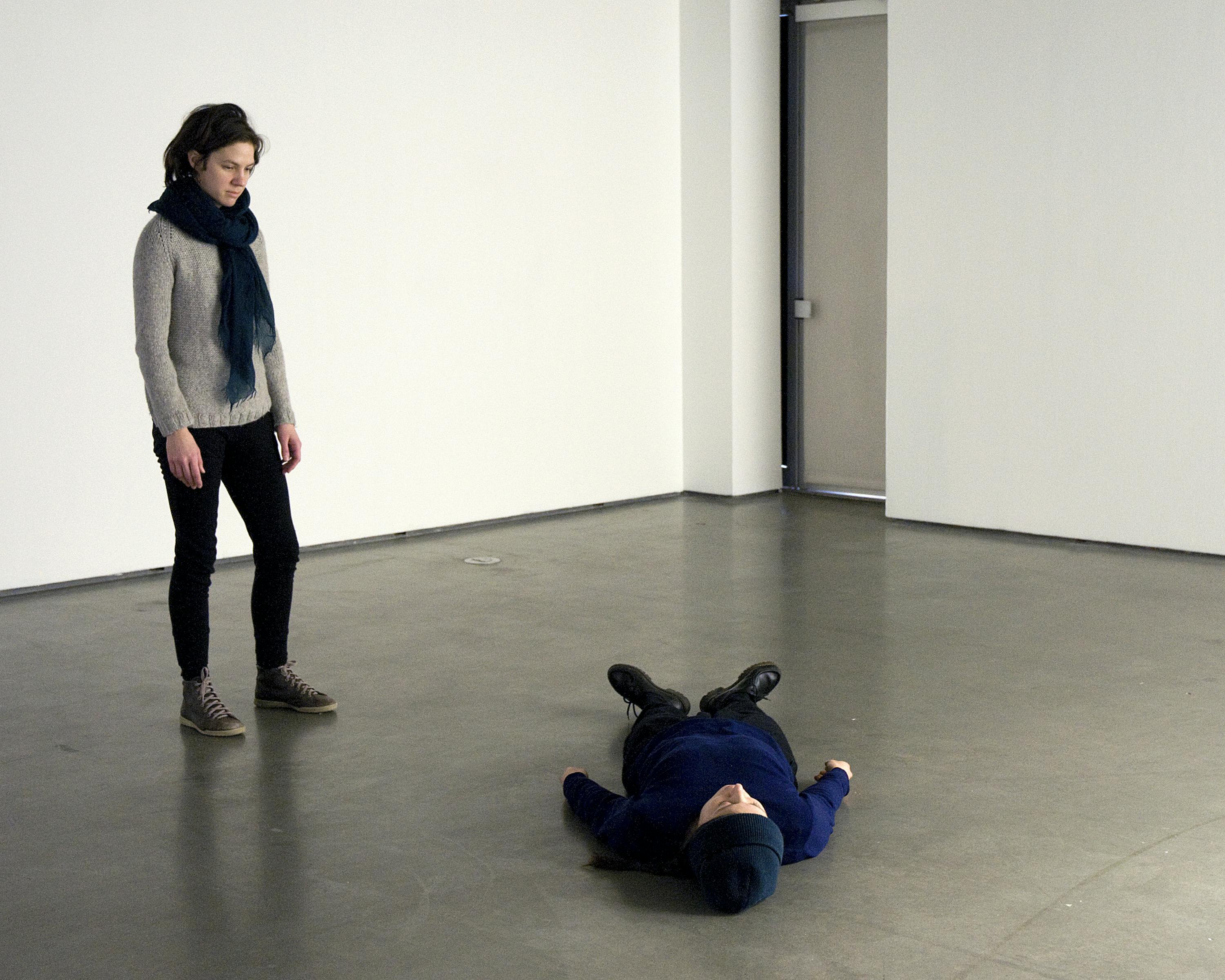 Two people are performing in a gallery space. The one wearing a blue-green scarf is standing straight and looking down at the other, who is lying down on the floor.
