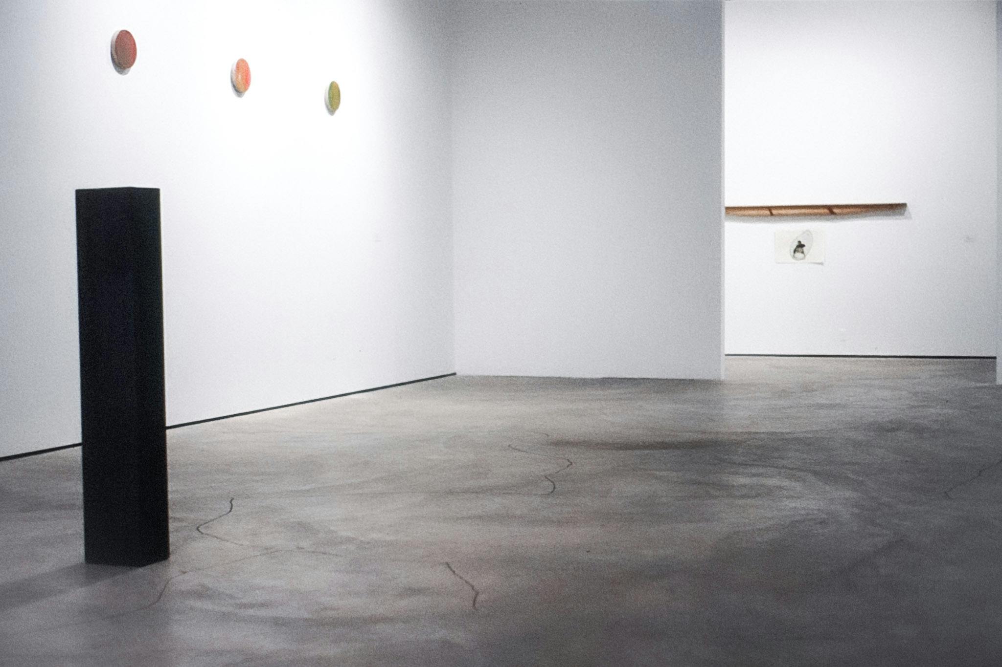 Three small circular artworks hang on a wall to the left, next two them is a tall black plinth and in the background part of another sculptural work is visible through a doorway. 