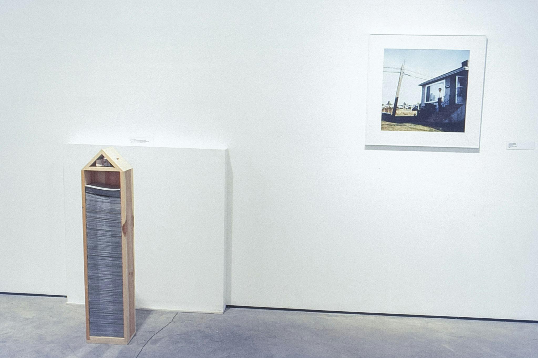 Two artworks in a gallery space. One is a small, vertical, house-shaped wood structure holding a tall stack of papers. The other is a colour photo of a person on the steps of a small grey house.