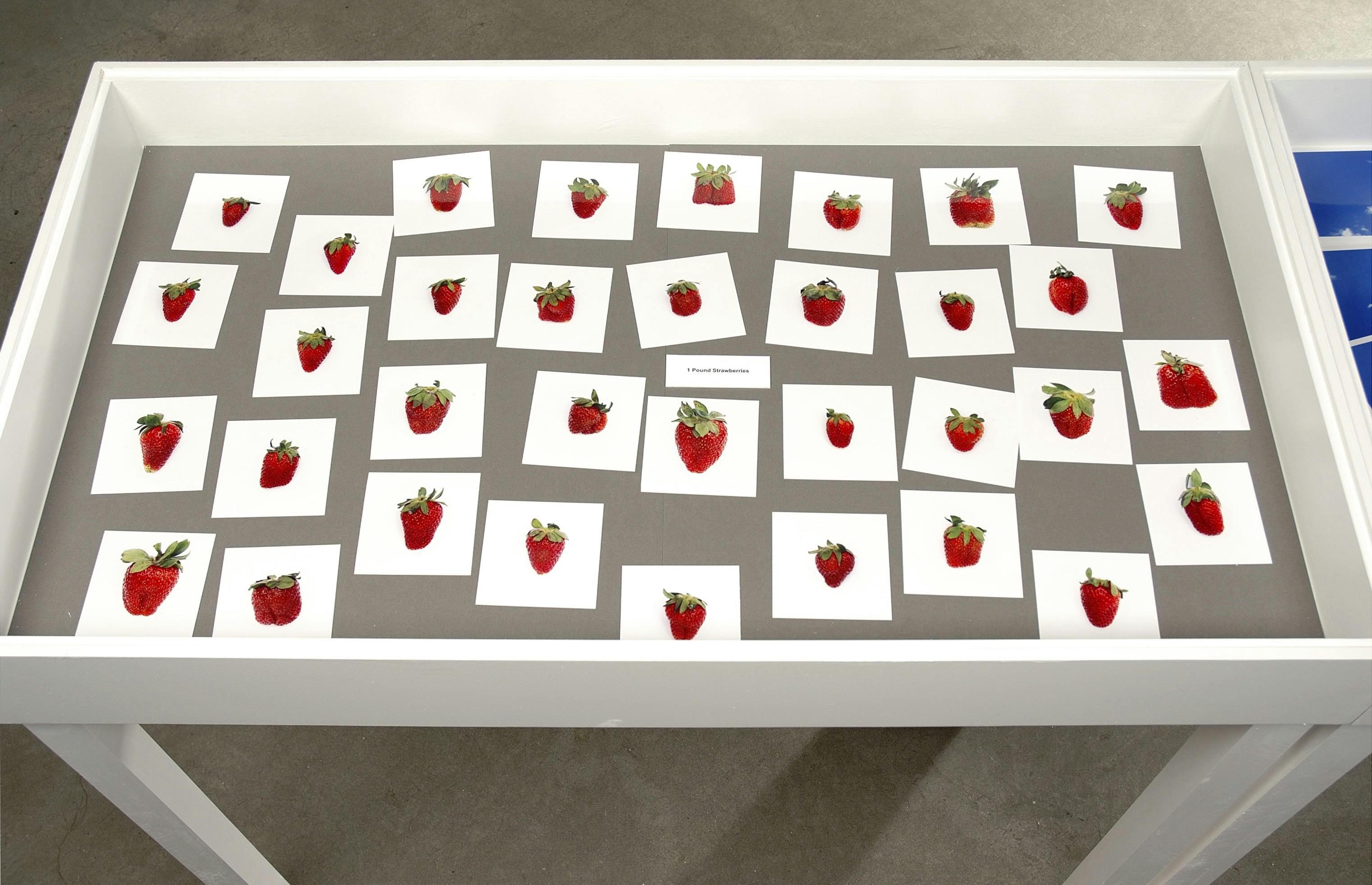 A closeup of a tabletop display. The main table in the photo shows several photos of strawberries of different sizes and shapes. Another table with images of a blue sky with clouds is partly visible.