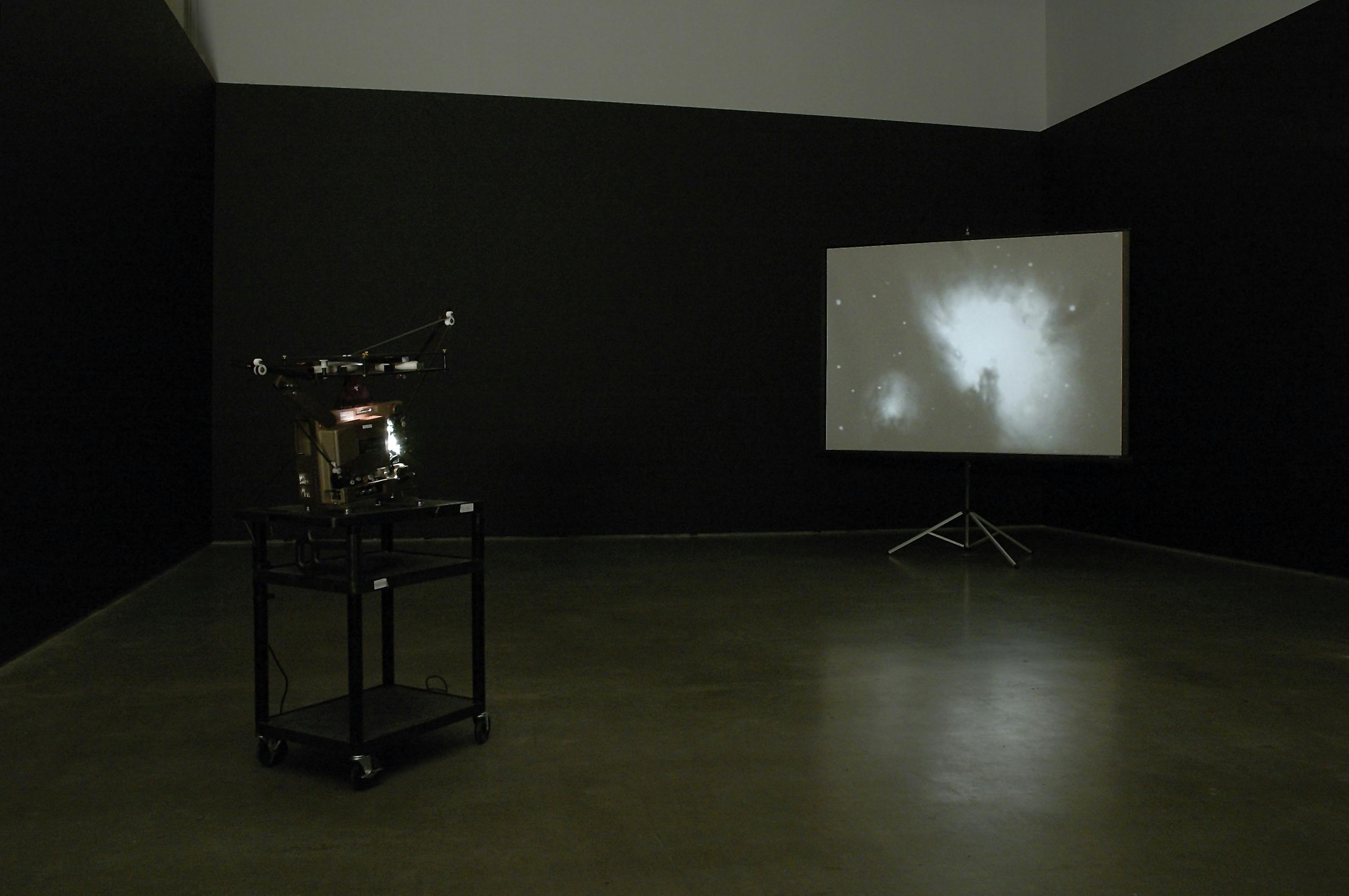 A film projector is installed in a darkened gallery space. A projector placed in a corner of the room shows a moving image of outer space. White-blue stars and a nebula are depicted in the image. 