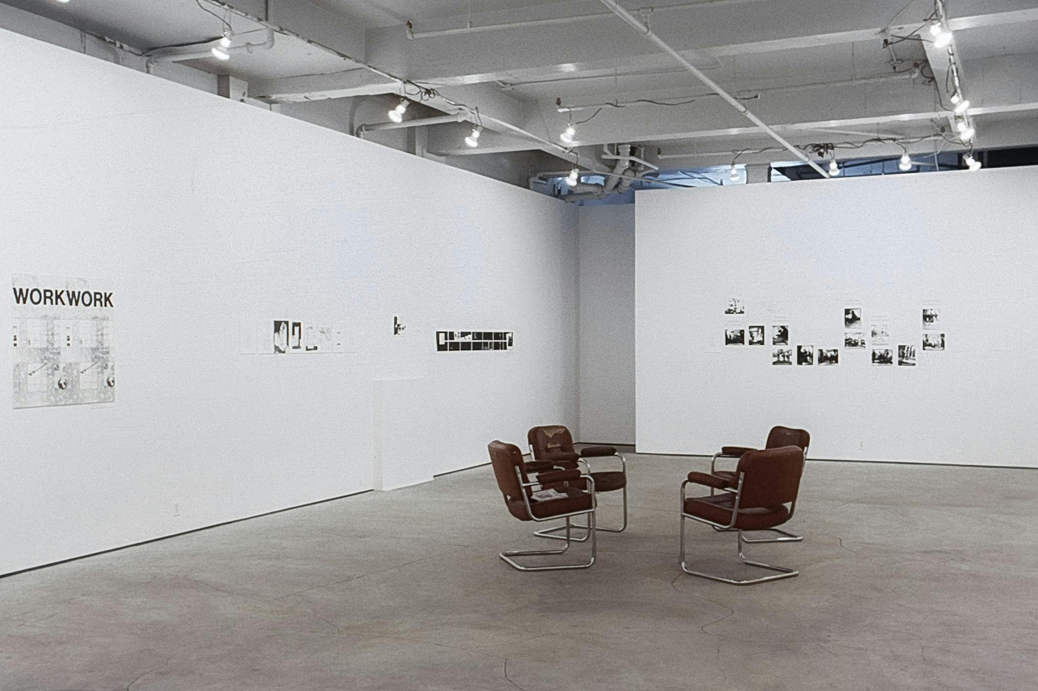 A gallery with artworks on the walls. Most are small black and white photos with text. One is a large set of blueprints, with letters reading "WorkWork." In the centre of the room, there are 4 chairs.
