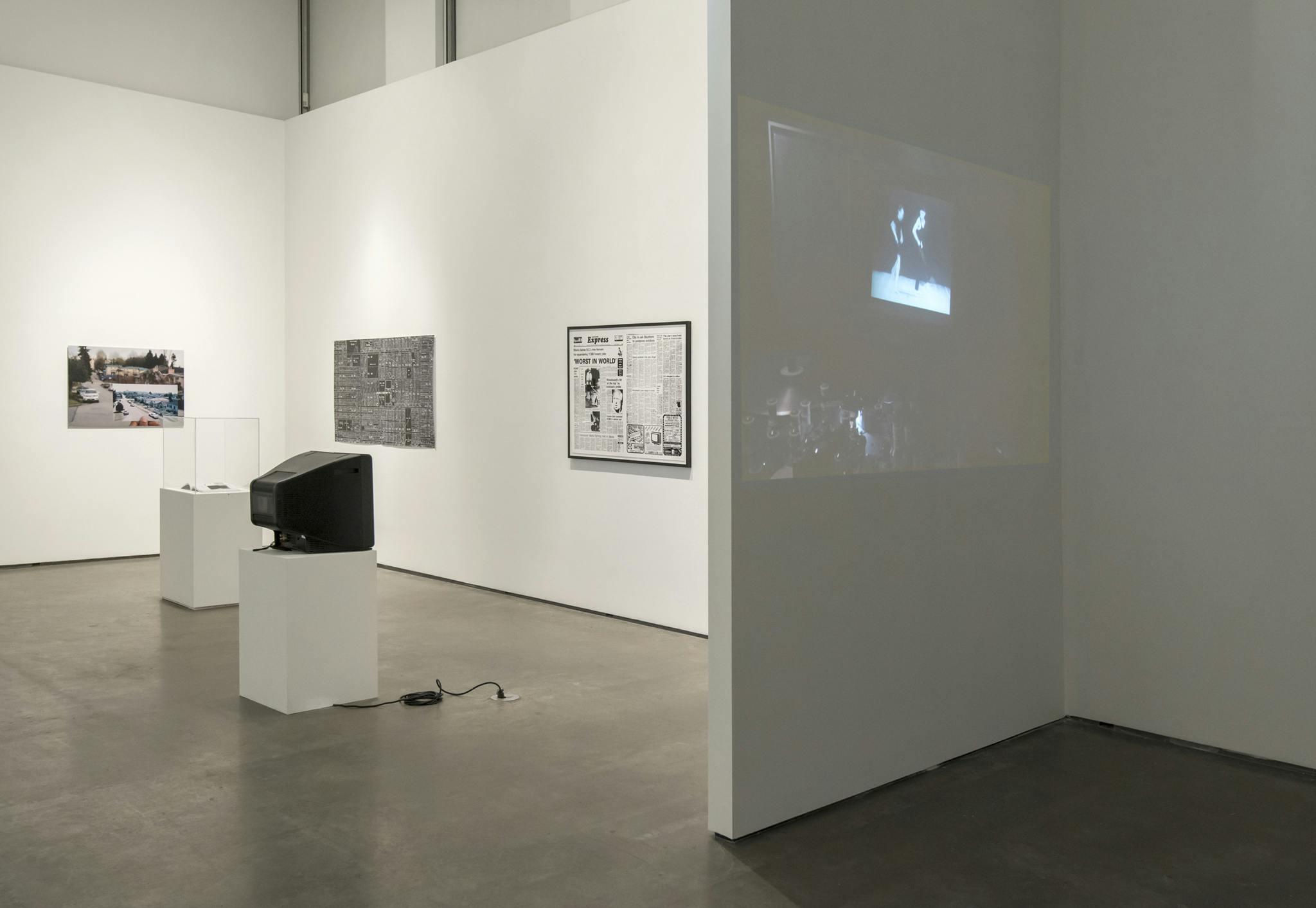 Multimedia works installed in a gallery: a video projected onto the white gallery wall, a TV monitor on a plinth, a framed print of a newspaper spread, a photograph, and a print of city subdivisions. 