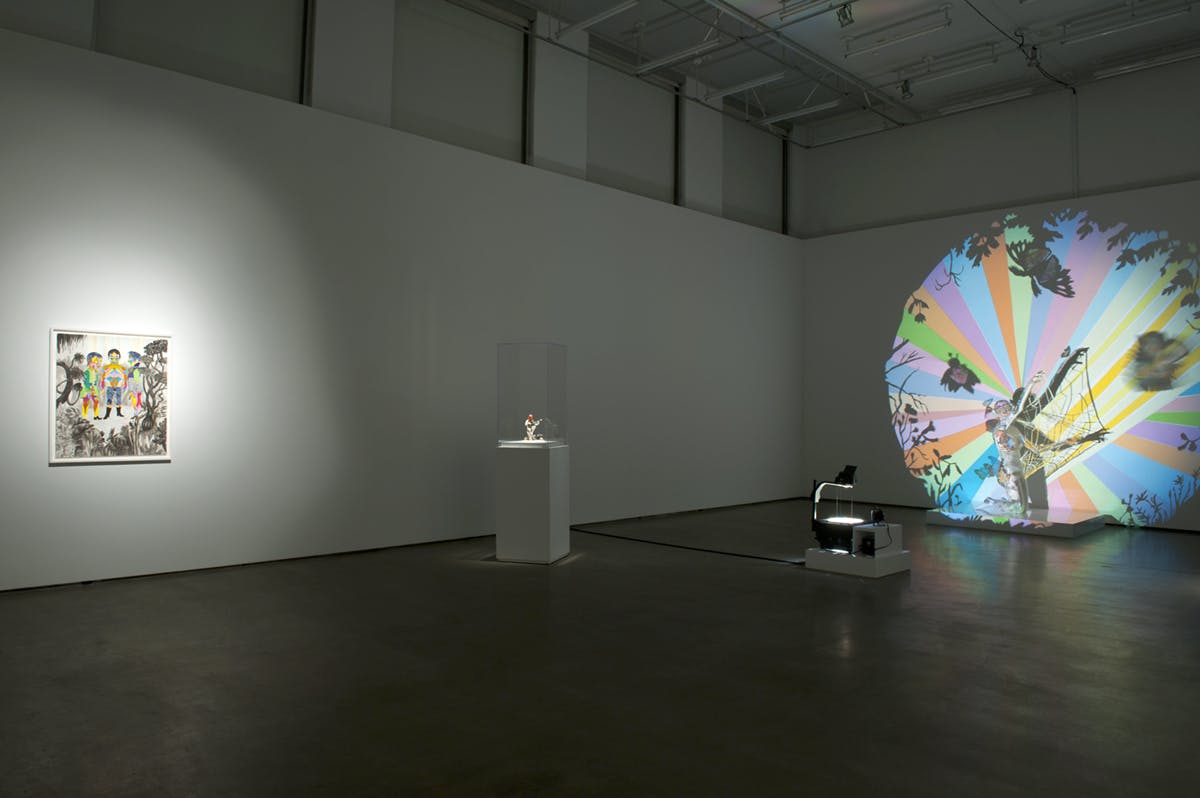 Three artworks are displayed in a gallery; a framed artwork on one wall, a small sculpture in a display case, and a colourful projection and sculpture on another wall. 
