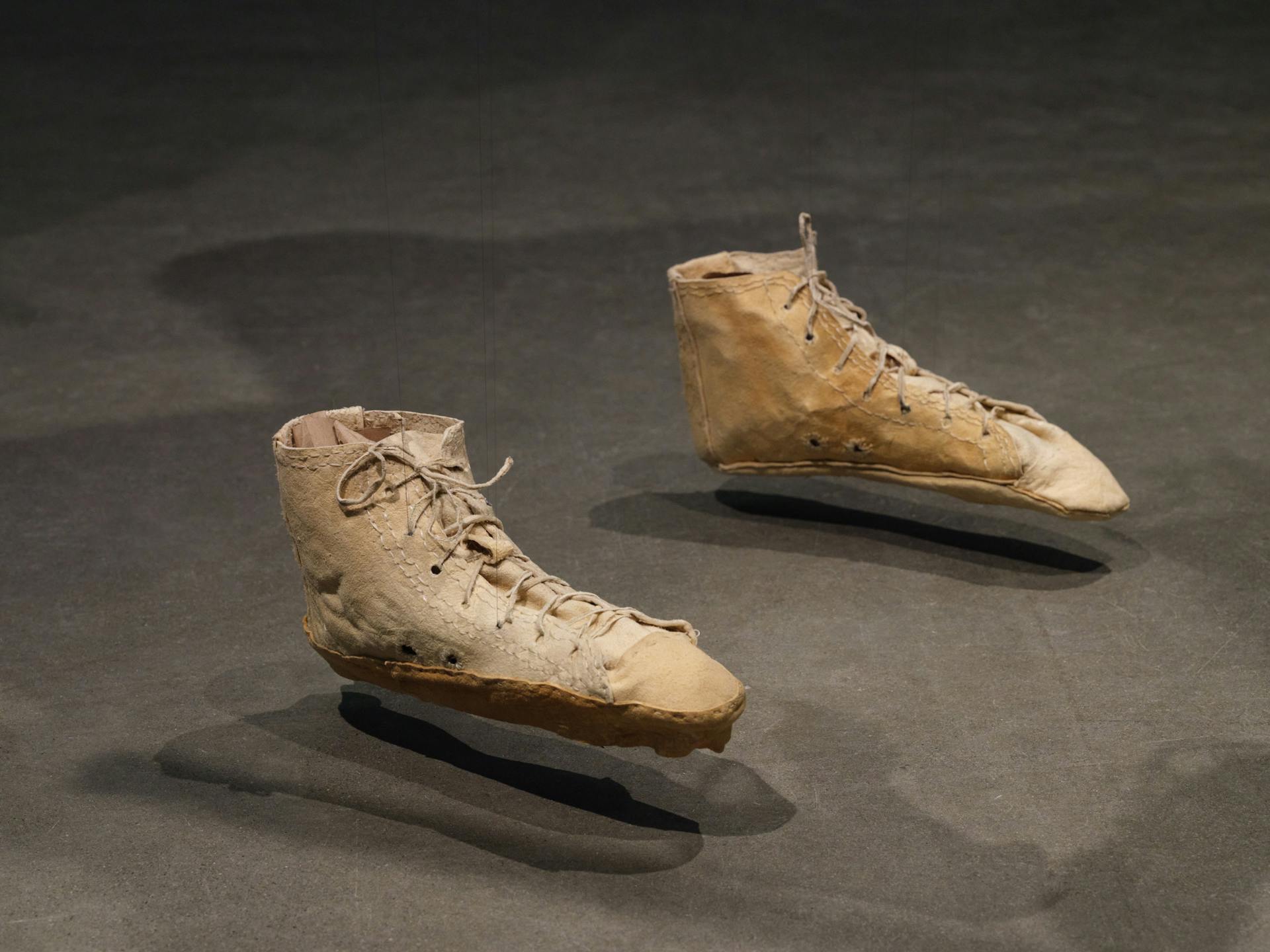 a pair of converse all-star-style sneakers made from tanned hide, suspended several inches above a concrete floor. the sneakers' soles resemble moccasins.