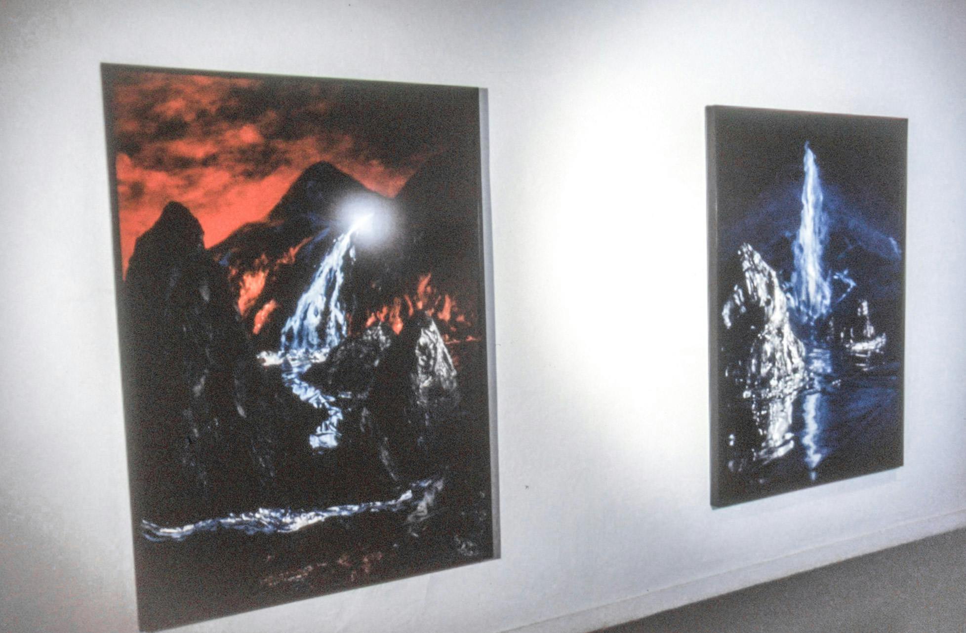2 very large photos mounted on the wall of a dark room. One photo appears to show an erupting volcano with a red sky, and the other shows a mountain-like glacier and water scene. 
