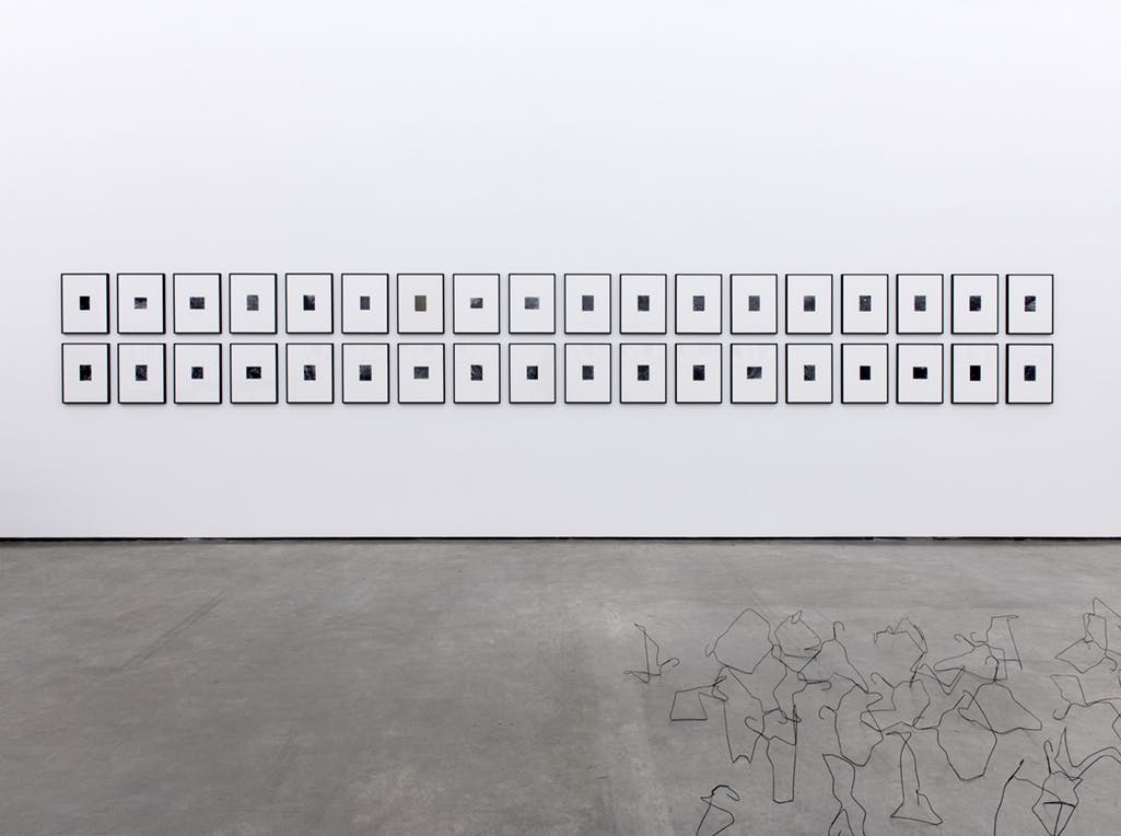 James Welling’s artworks installed in a gallery. In this image, 36 small black and white images, each framed, are mounted on the wall. Several black wire sculptures sit on the floor in front of the wall.