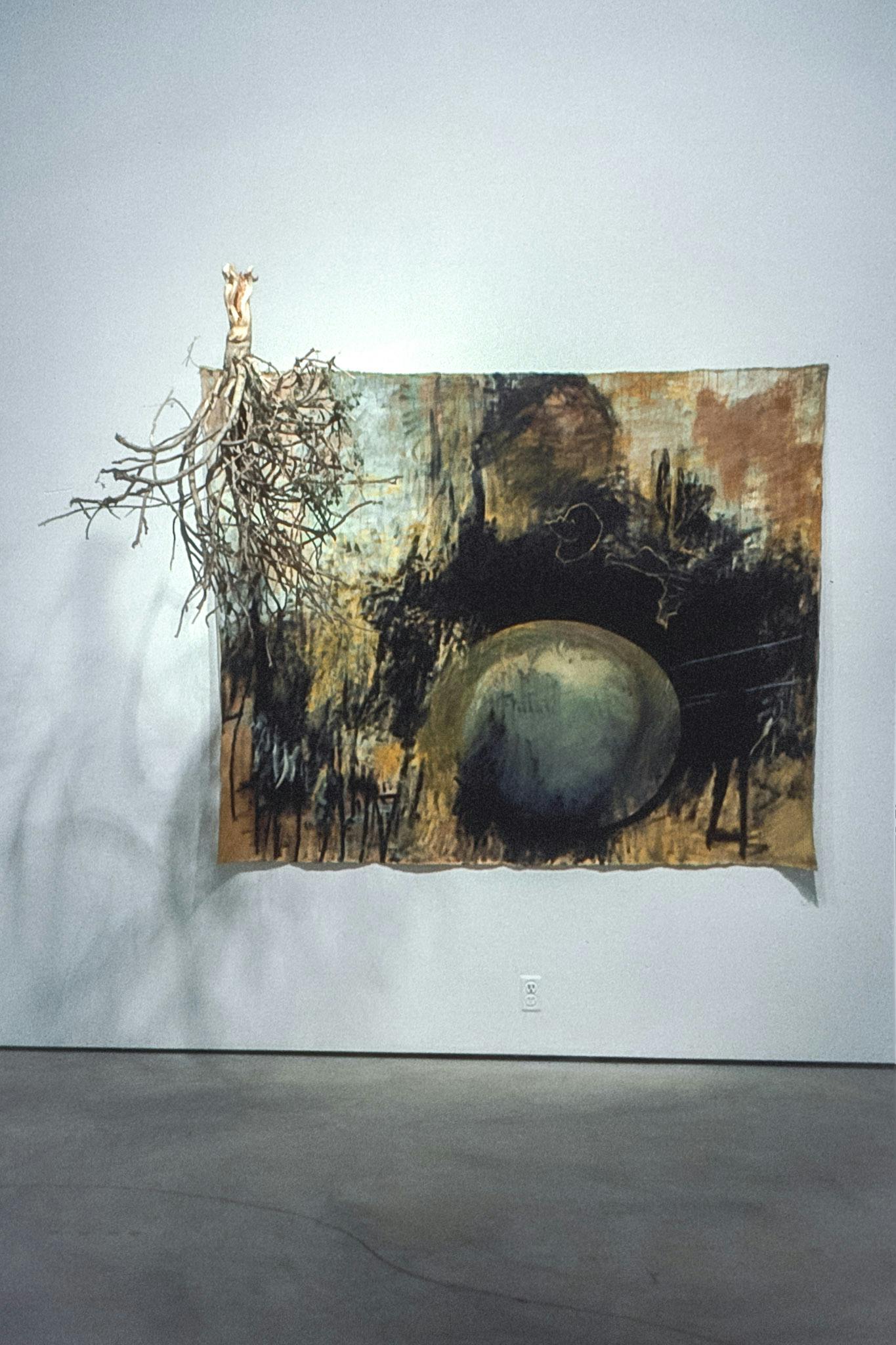An artwork on a white wall. The work is a painting on fabric pinned to the wall, containing rusty, earthy tones. Extruding from the painting is a large wood branch, possible with tree roots attached.