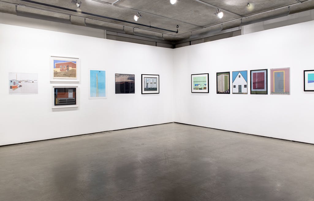 Multiple drawings by Itee Pootoogook hang on the walls of a gallery. In total, twelve framed drawings are visible in this photo. Several of these drawings show buildings standing in natural landscapes.