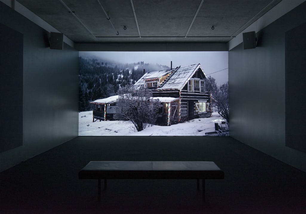 Video projection on a dark gallery wall of a house made of log timber. The house sits in a snowy landscape with forested hills behind. The outdoor light is faint, it's either dusk or dawn. 