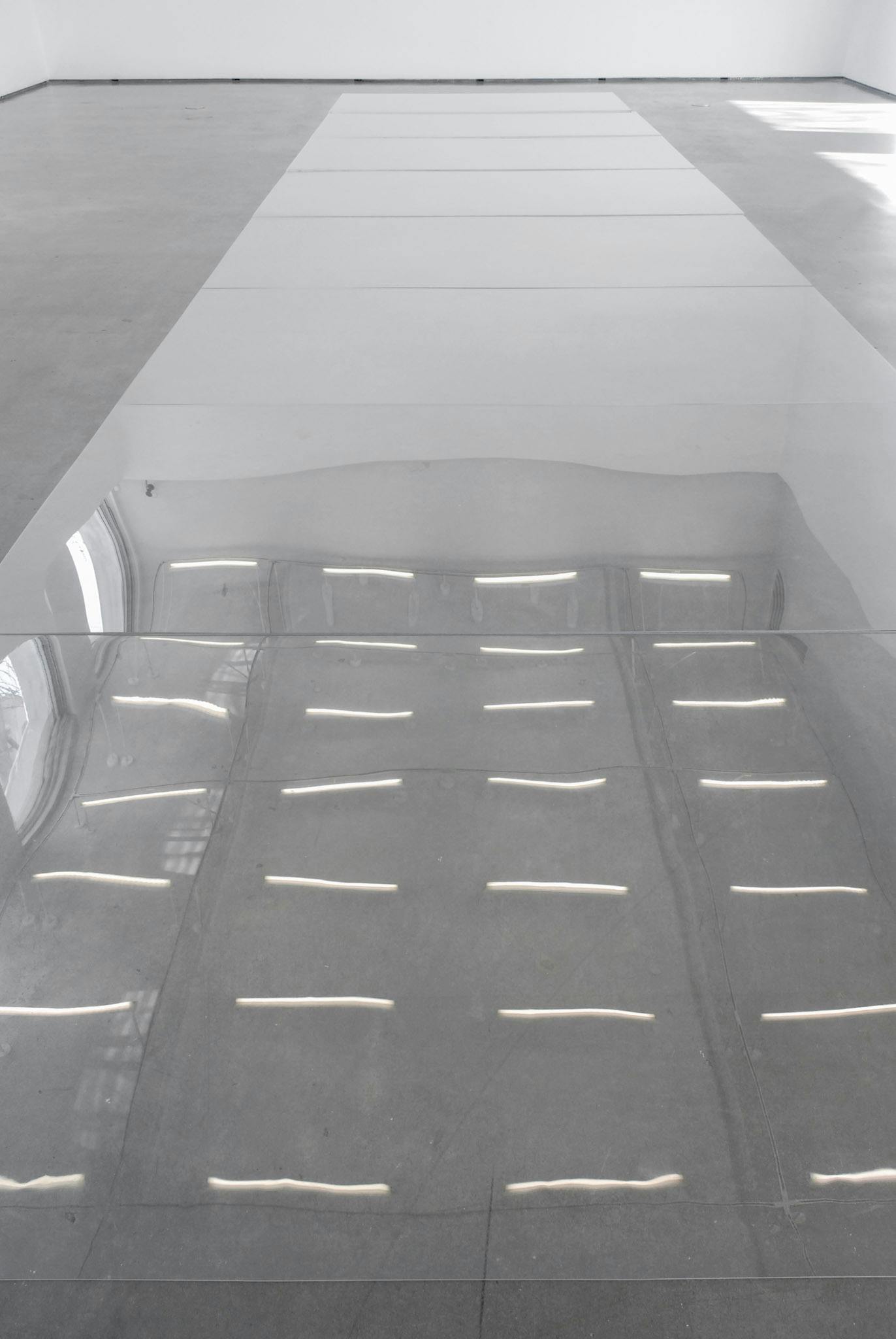 A number of rectangular-shaped mirrors are placed in a row on the gallery floor. This row of mirrors reflects the white gallery ceiling, on which many white fluorescent lights are installed.