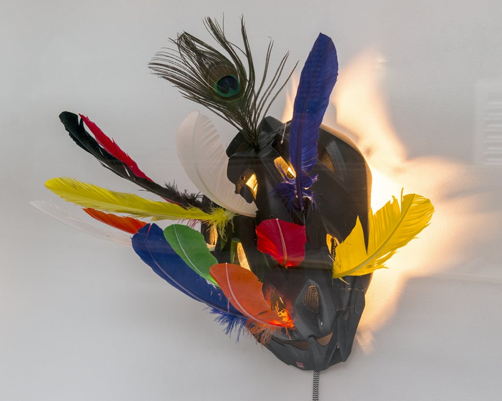Detail image of a sculpture by Ryan Gander. A black bike helmet, affixed with colourfully dyed feathers, is illuminated by a yellow light placed inside the helmet. 