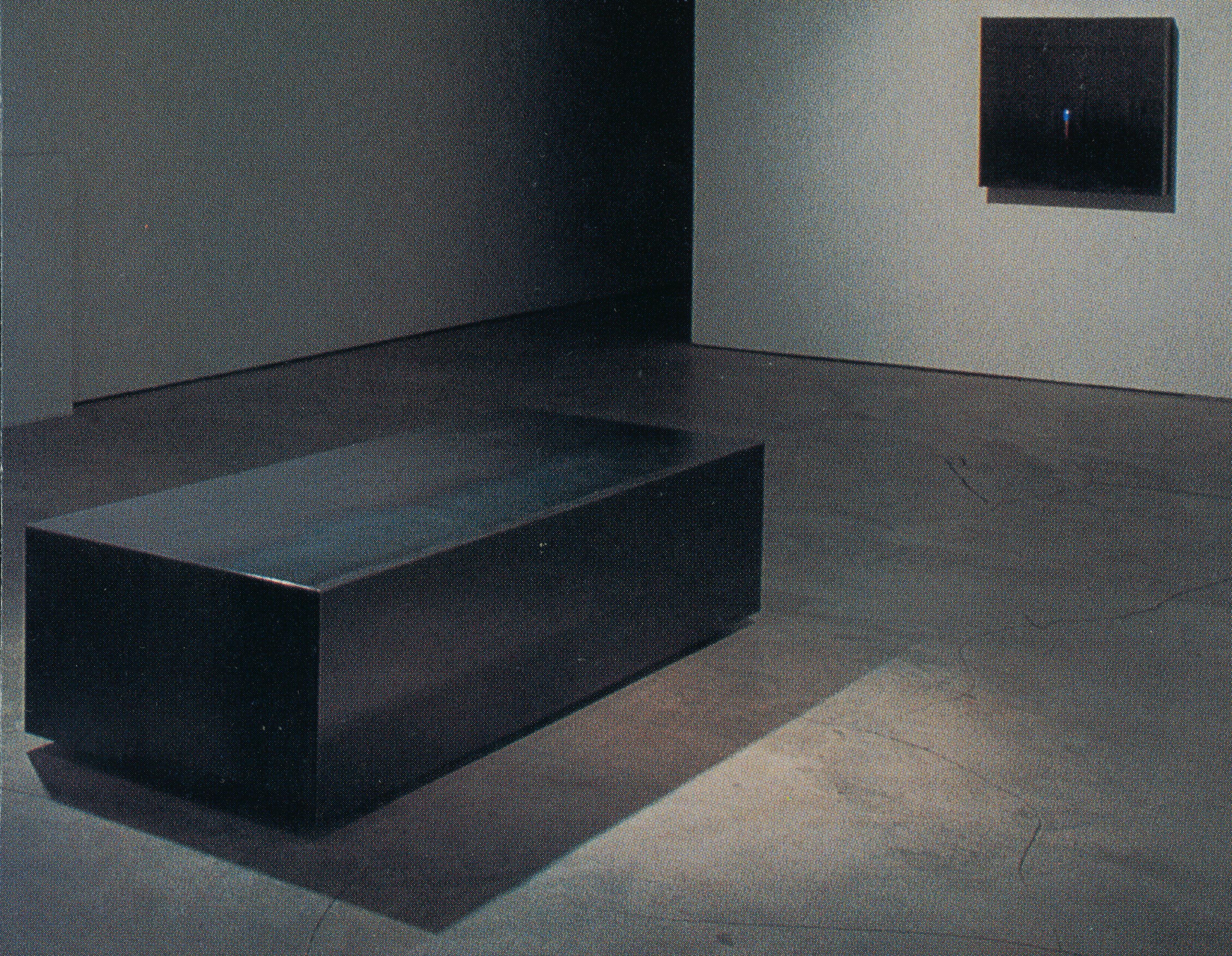 A large steel-made sculpture is placed in the middle of the dimly lit gallery. It is rectangular and has a tarnished dark grey colour. The sculpture is directly placed on the floor. 