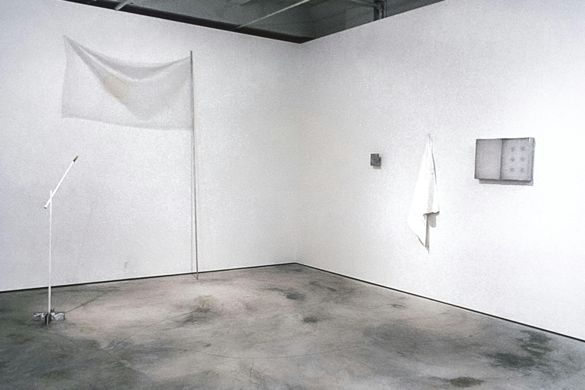 Several objects in the corner of a gallery. The objects include a large white flag leaning on the wall, two small paintings on the wall, and a white construction that resembles a microphone stand. 