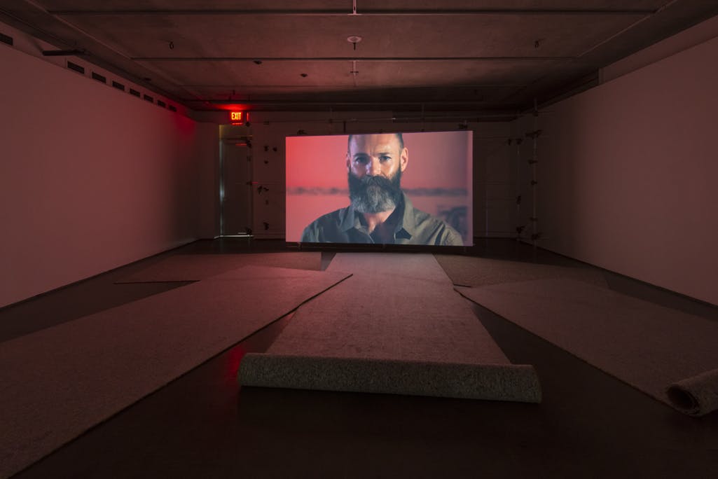 A large screen is installed at the end of the gallery space, on which a bearded face is projected. Dim red light illuminates the room. Five long carpets are placed on the floor.