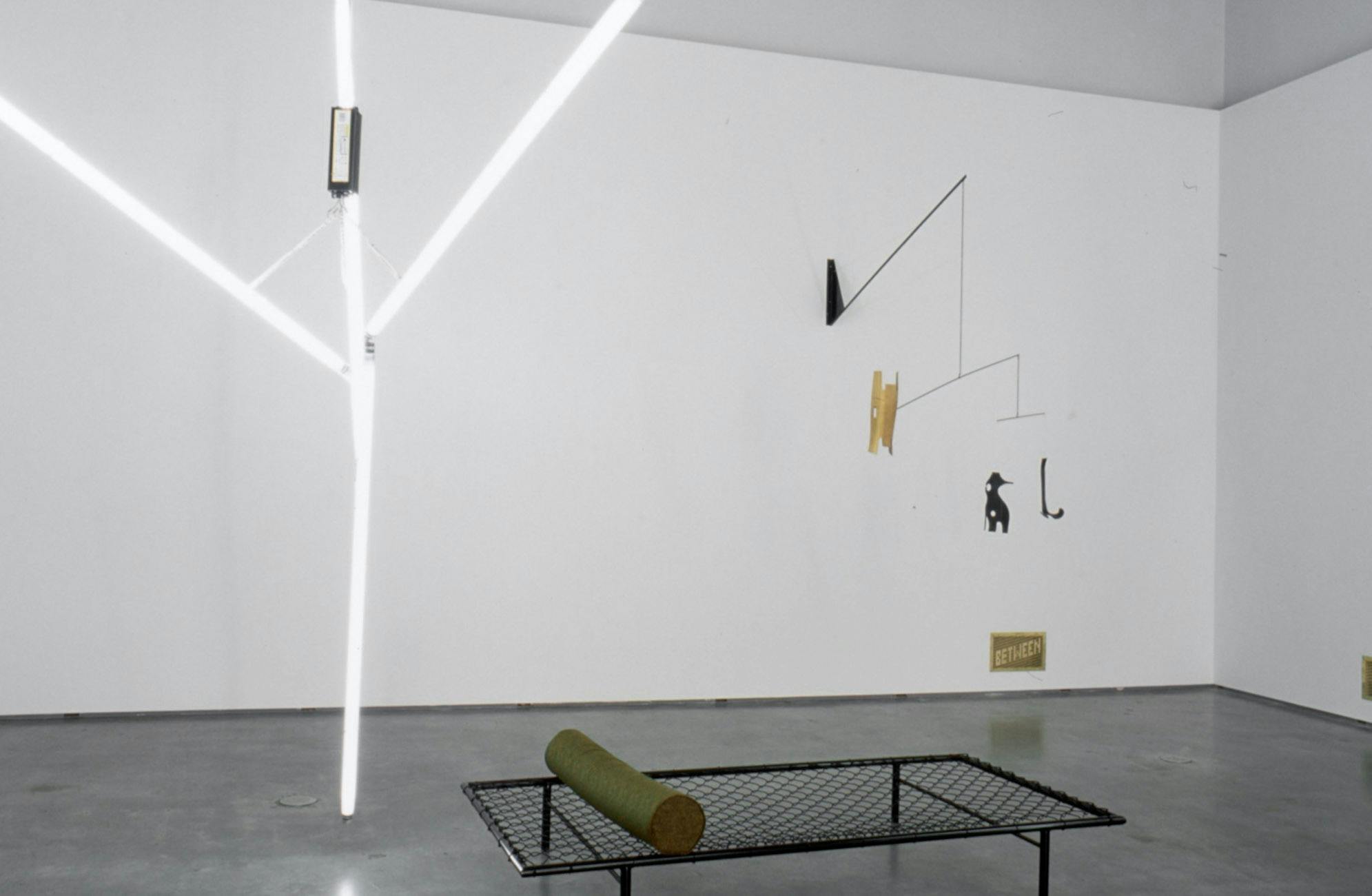 Three sculptures are installed in a gallery space. They are a large-sized sculpture made of tube light bulbs, a black bed frame, and a wire sculpture that hangs down objects to the air.