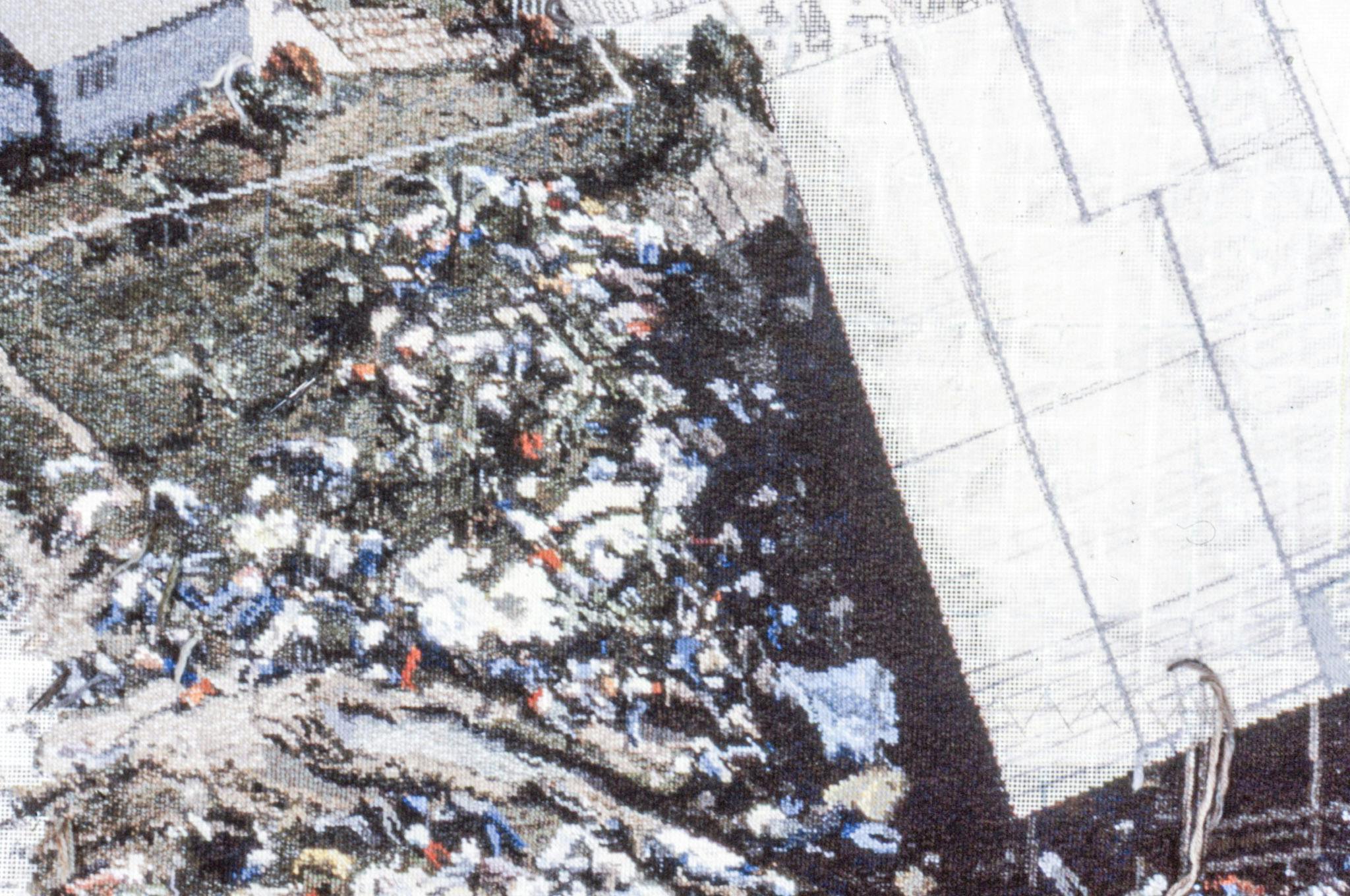 A detailed image of an embroidered carpet, with threads hanging from it. The carpet is based on a bird's-eye image of the Jonestown massacre, and this closeup shows a section with bodies on the grass. 