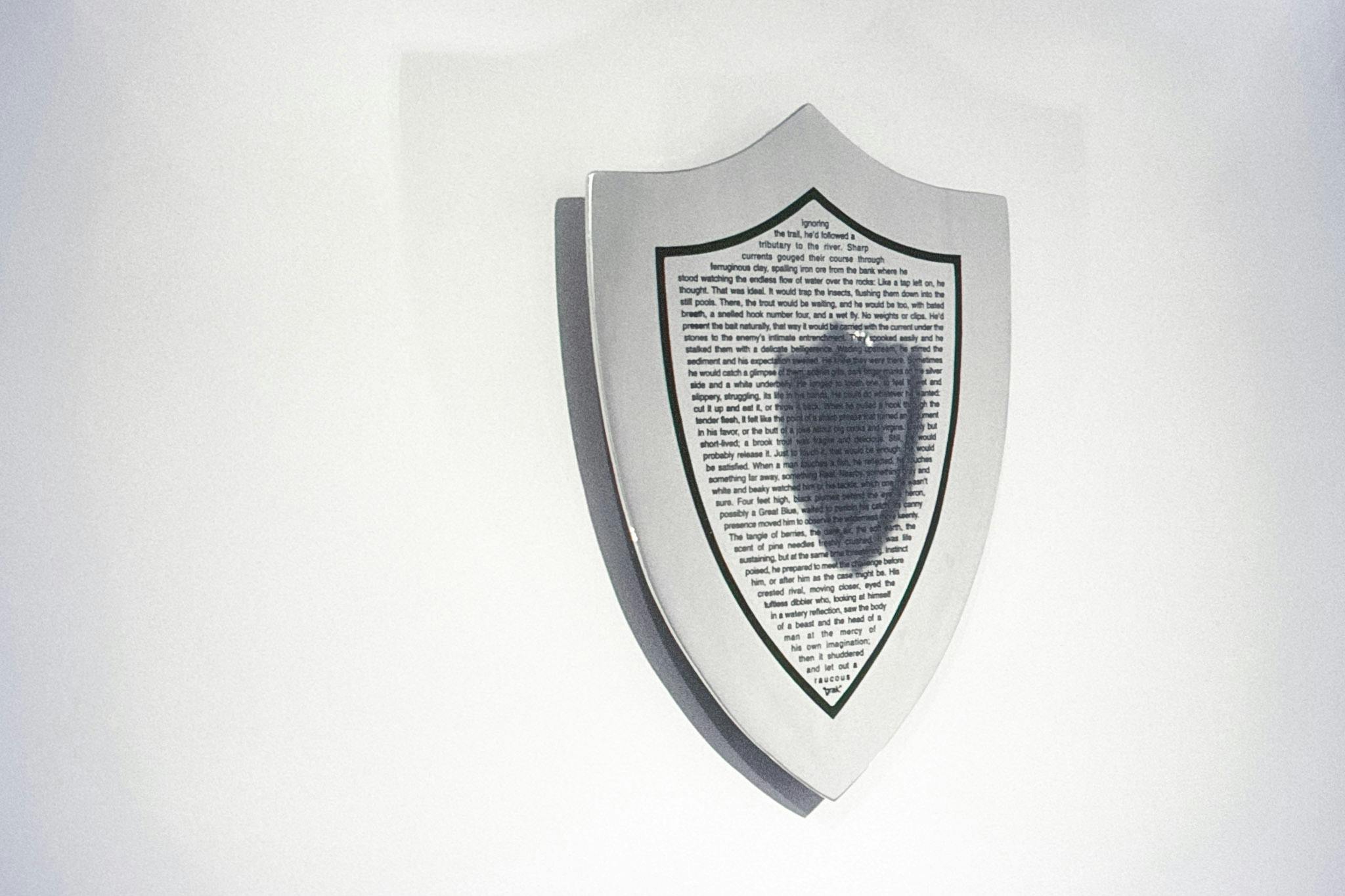 A closeup of an artwork on a white wall. It is a glossy silver shield with a black outline and small black text. The first sentence reads: "Ignoring the trail, he'd followed a tributary to the river."