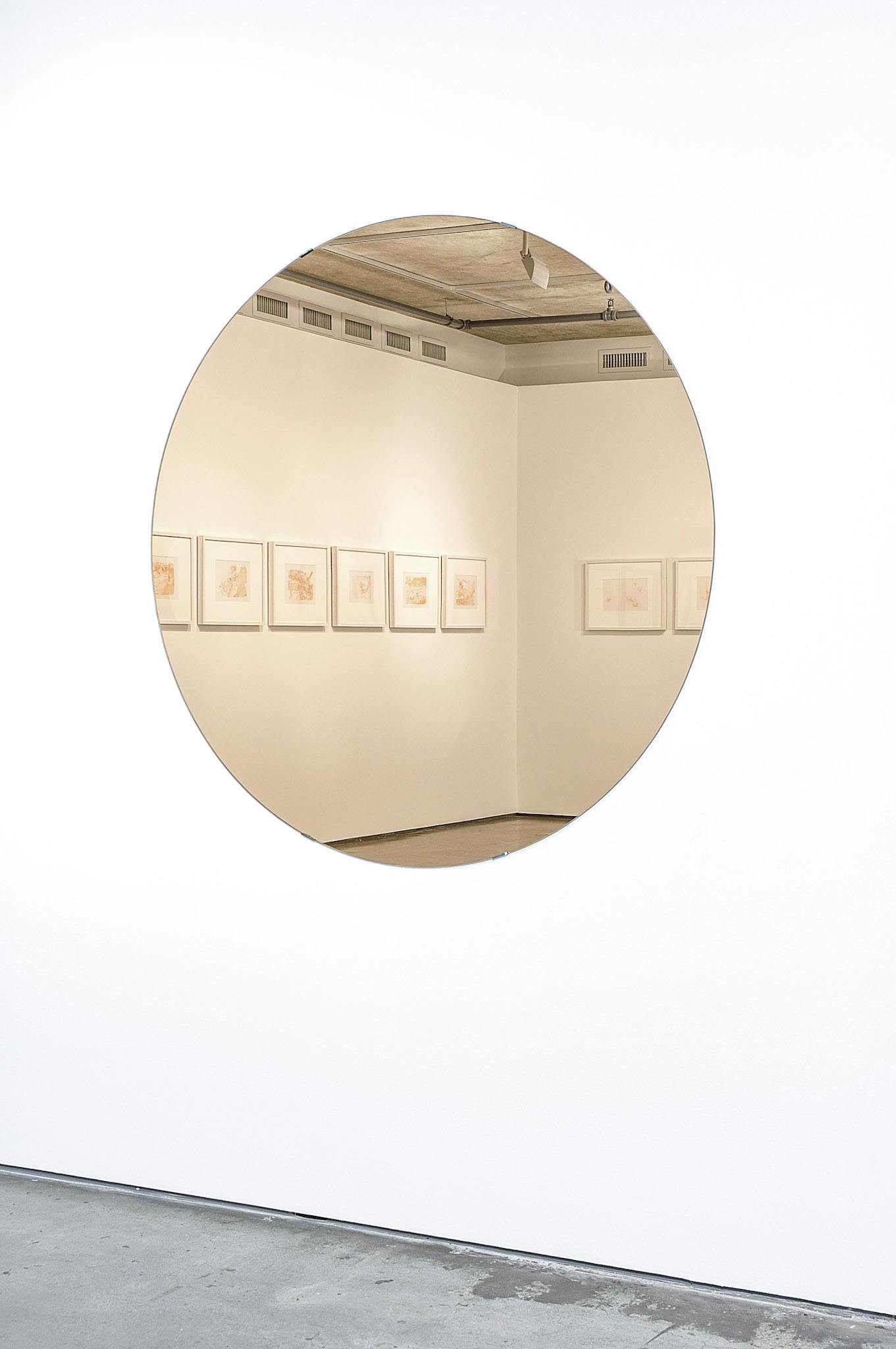  A closeup of a circular mirror mounted on a white wall. The reflection in the mirror shows the corner of a gallery, with several orange drawings in white frames.
