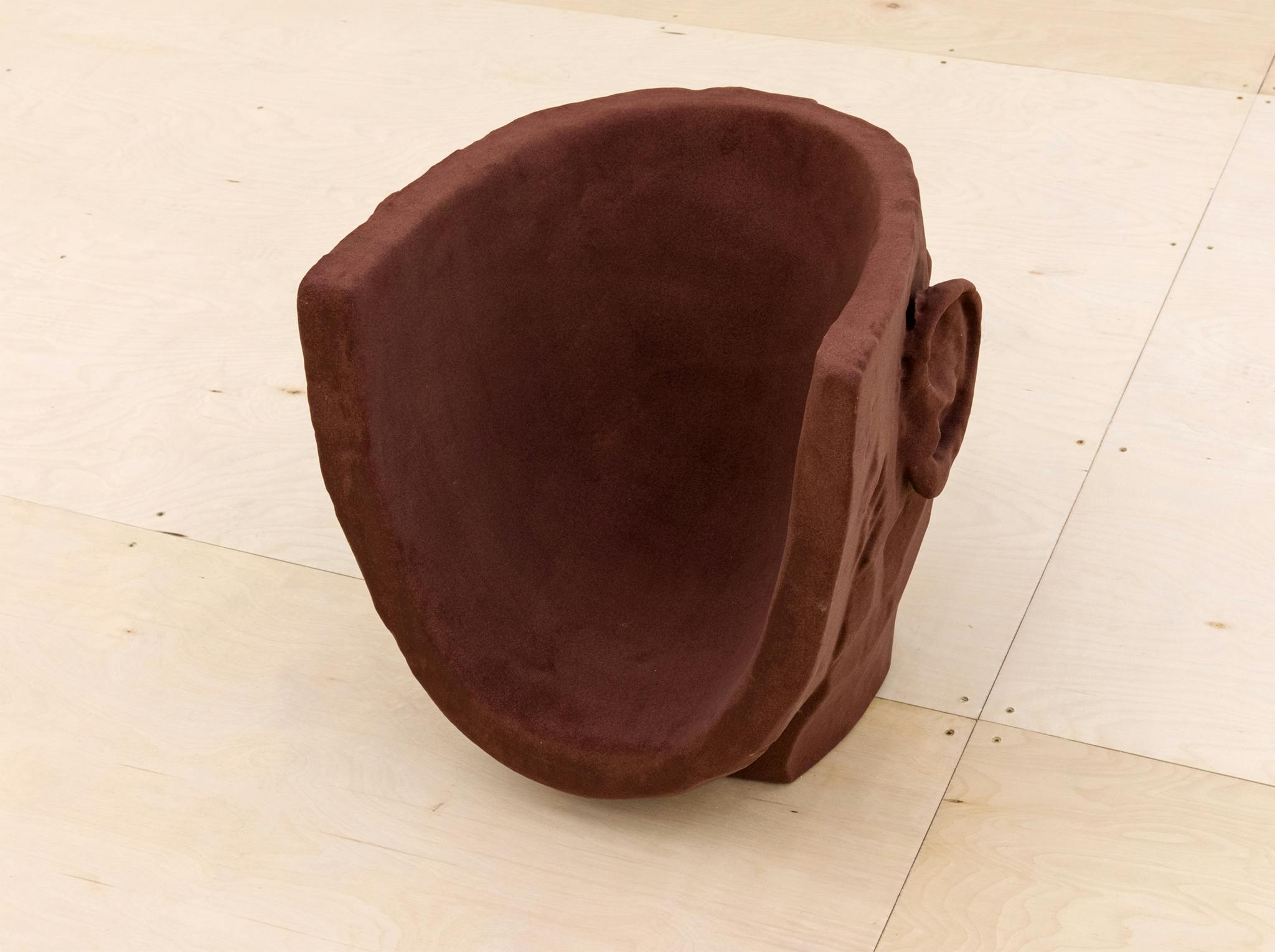 A brown sculpture resembling a human head is installed on a gallery floor. It is faceless, and hollowed out, making the sculpture look like a low chair. 