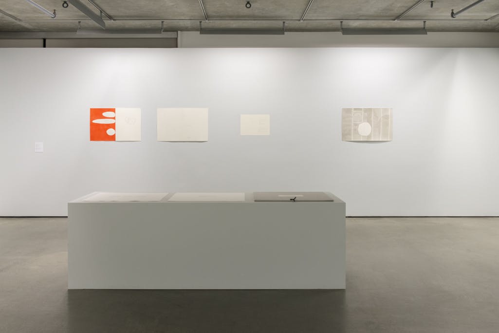 Multiple two-dimensional artworks are installed in a gallery space, including drawings and watercolour paintings. Several pieces are placed on a large plinth in front of a wall. 