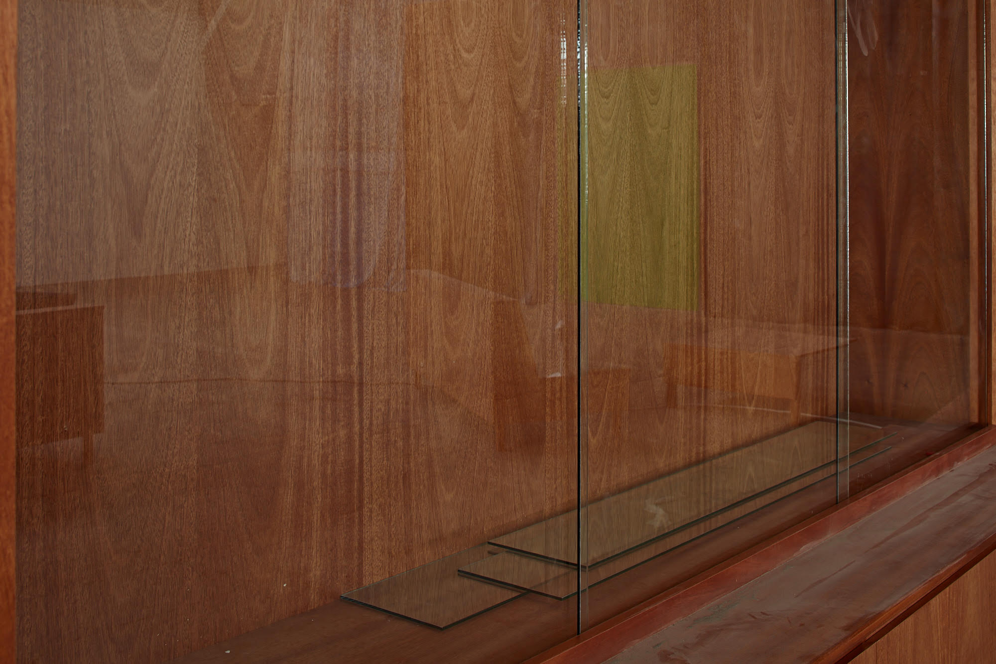 A closeup shot of a wooden cabinet with glass doors. A faint reflection of the surrounding room is visible in the glass, including scattered furniture and a doorway rendered in green paint.