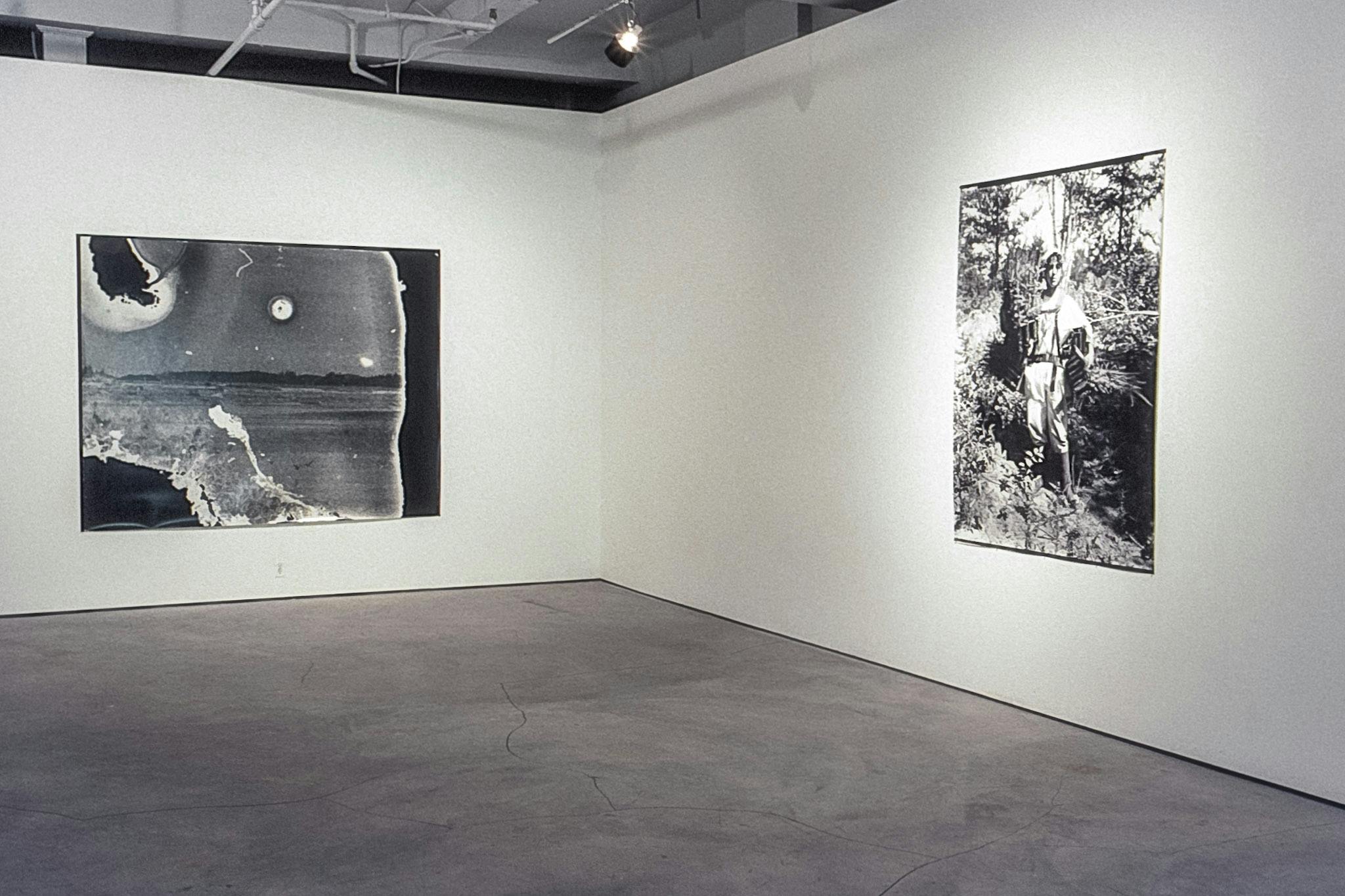 The corner of a gallery with 2 large black and white photos on the wall. The photo on the left shows a faint landscape on damaged film. The photo on the right shows a person standing in the woods.