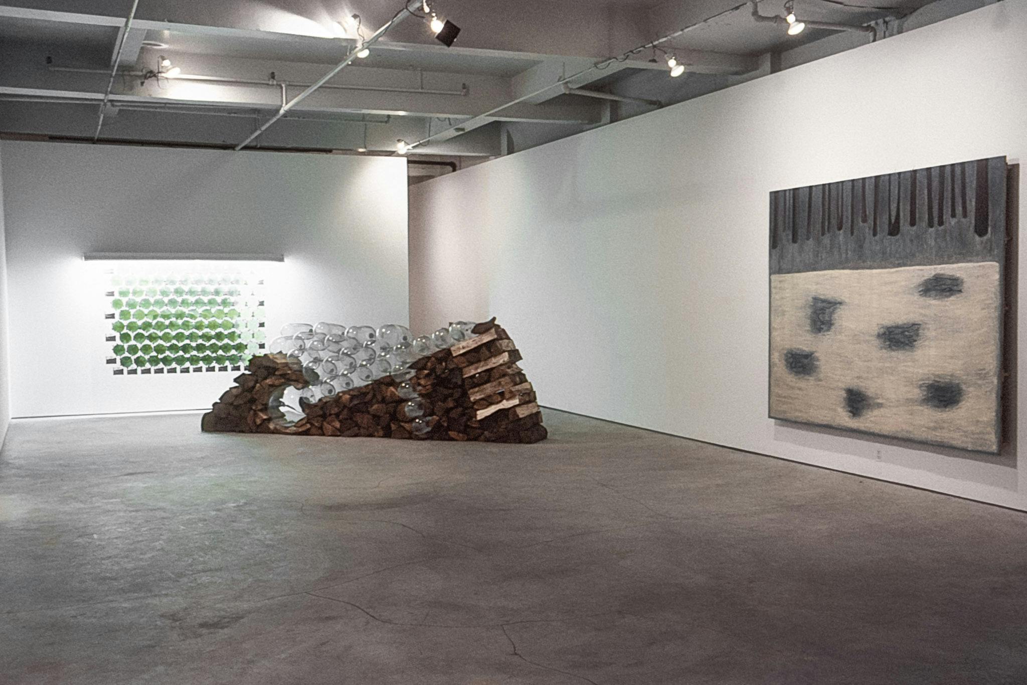 A gallery with 3 visible artworks. One is an illuminated display of green and black shapes on the wall, one is a stack of firewood and glass cylinders, and one is a gray, black, and yellow painting.