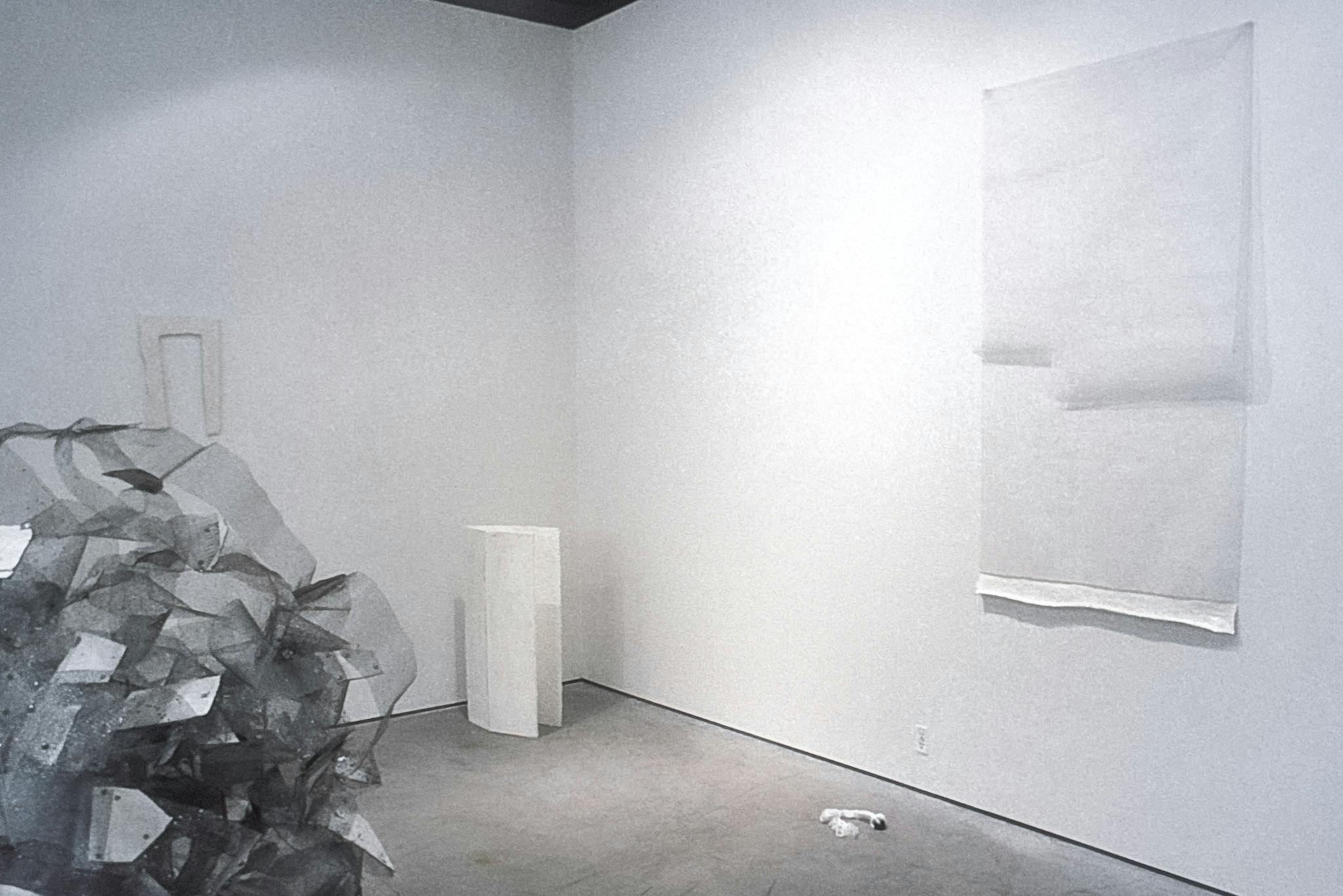 A closeup of artworks in a gallery. The works include sheer white sheets on the wall, a small textile object on the floor, and a tall and white box, with open flaps.