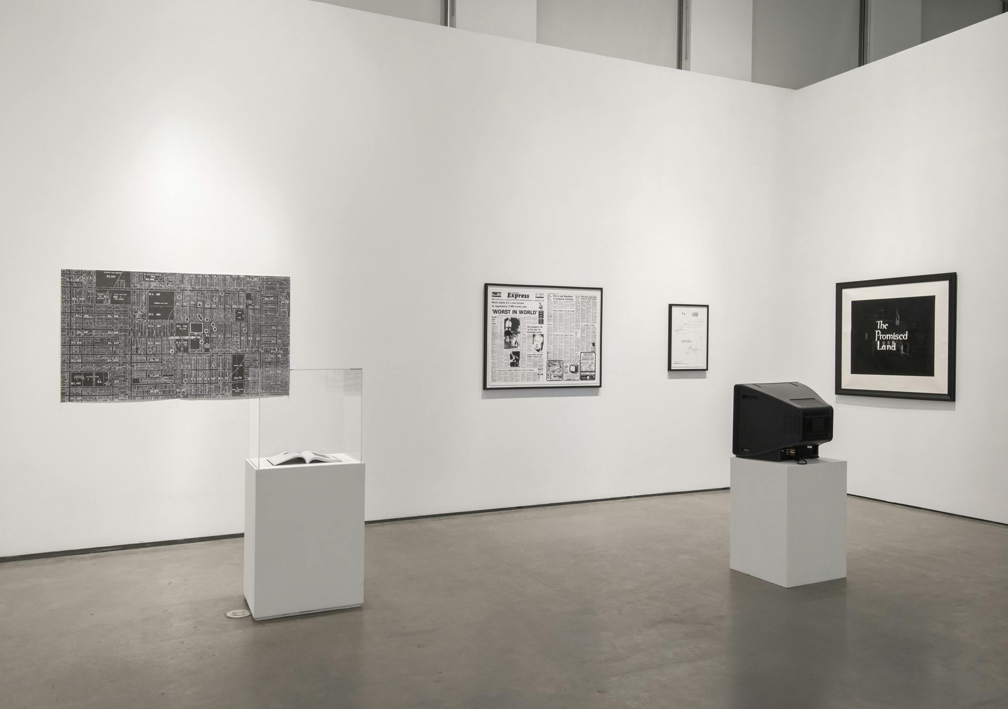 Multimedia works installed in a gallery: a TV monitor on a plinth, two framed prints, a framed print of a newspaper spread, a print of city subdivisions, a vitrine with an open book.