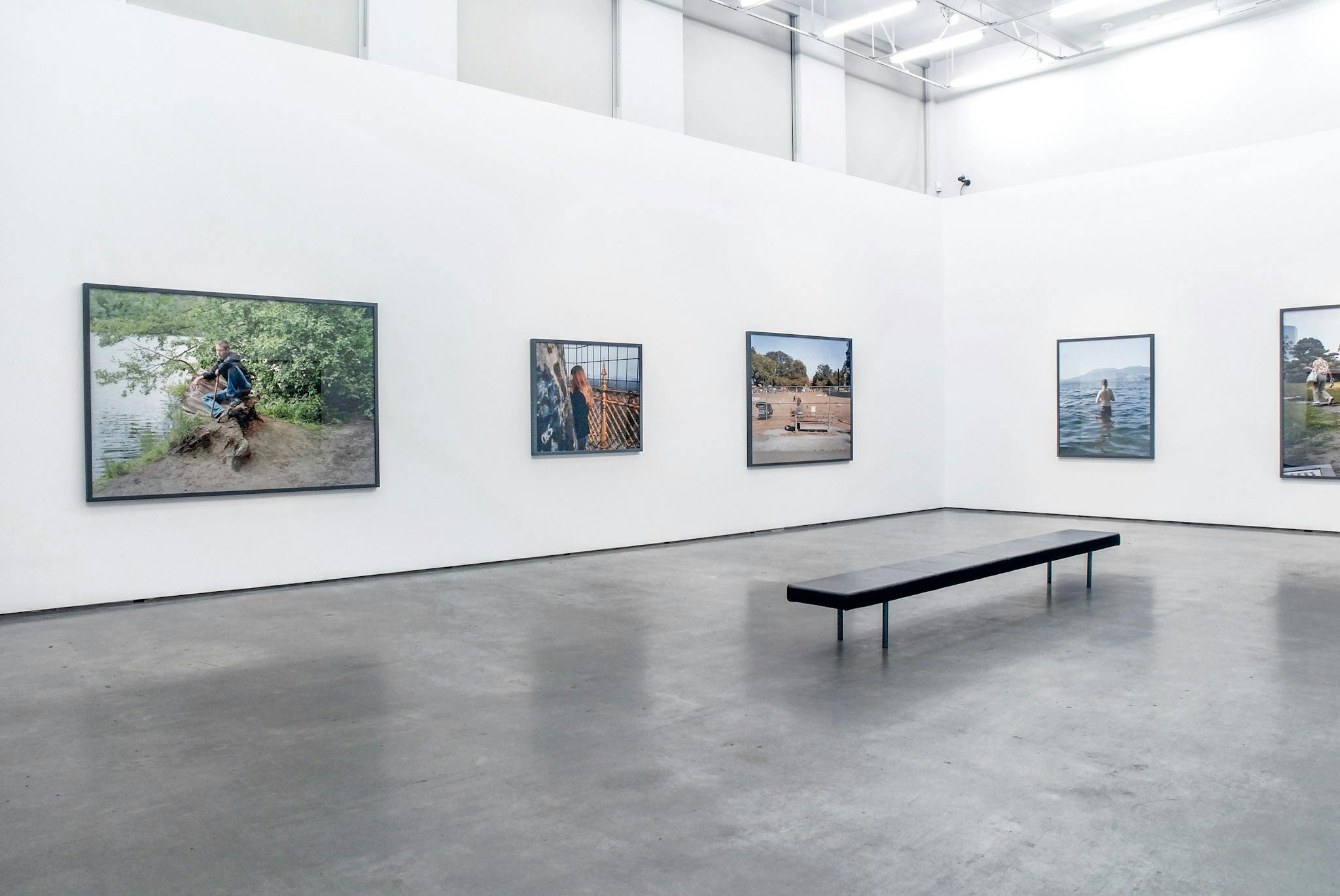 Various large-sized coloured photographs are installed in a gallery space. The photograph on the left depicts a young person sitting on a tree stump beside a body of water. 