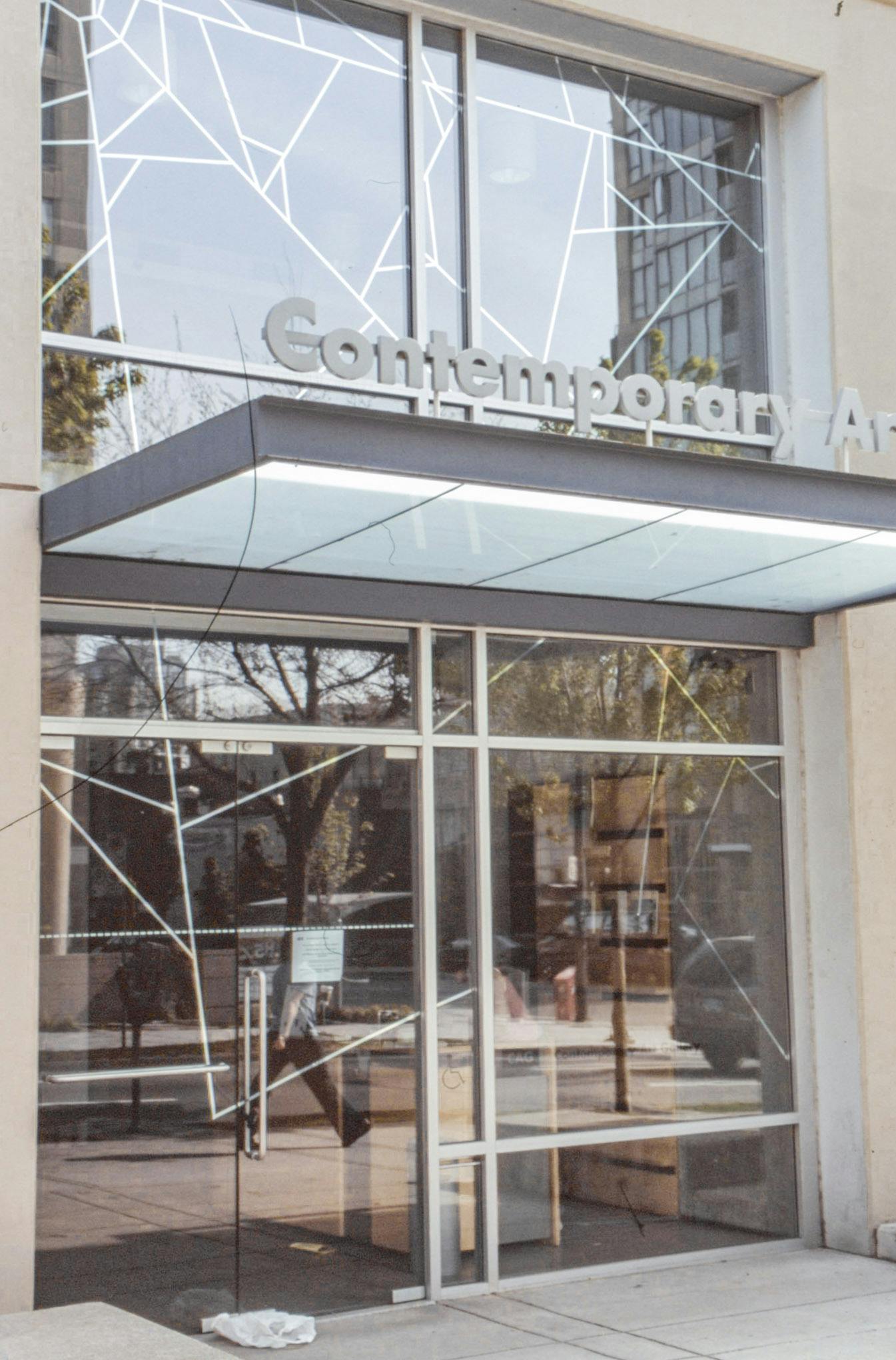 An image showing the front entrance door of the CAG seen from outside. A vinyl of a white spider’s web is printed on the CAG’s glass front door and windows above and besides it.