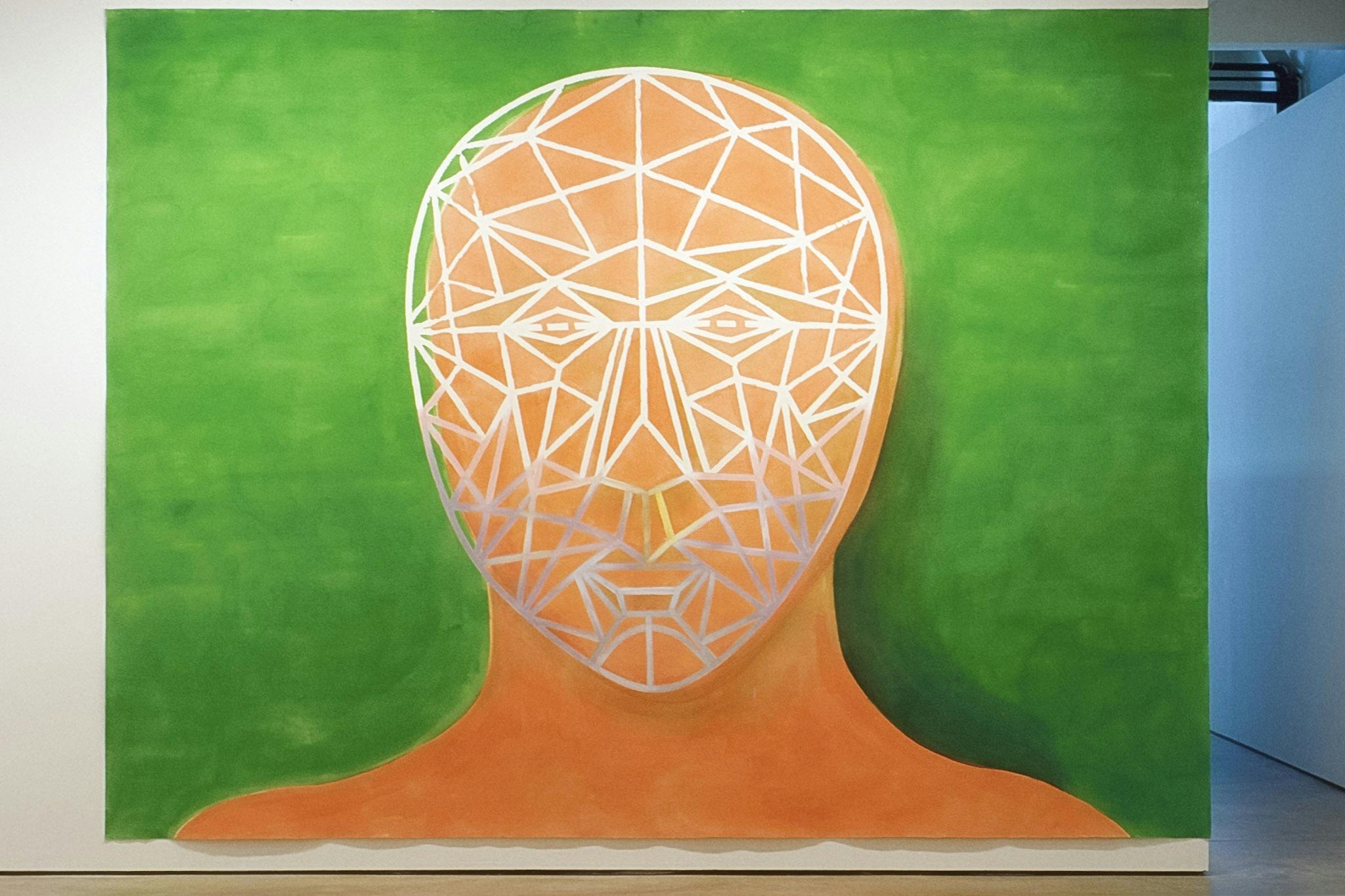 An unframed large painting is installed on the gallery wall. An orange human head is depicted in the green background. A human face, made of a white web, is drawn over the faceless orange head. 
