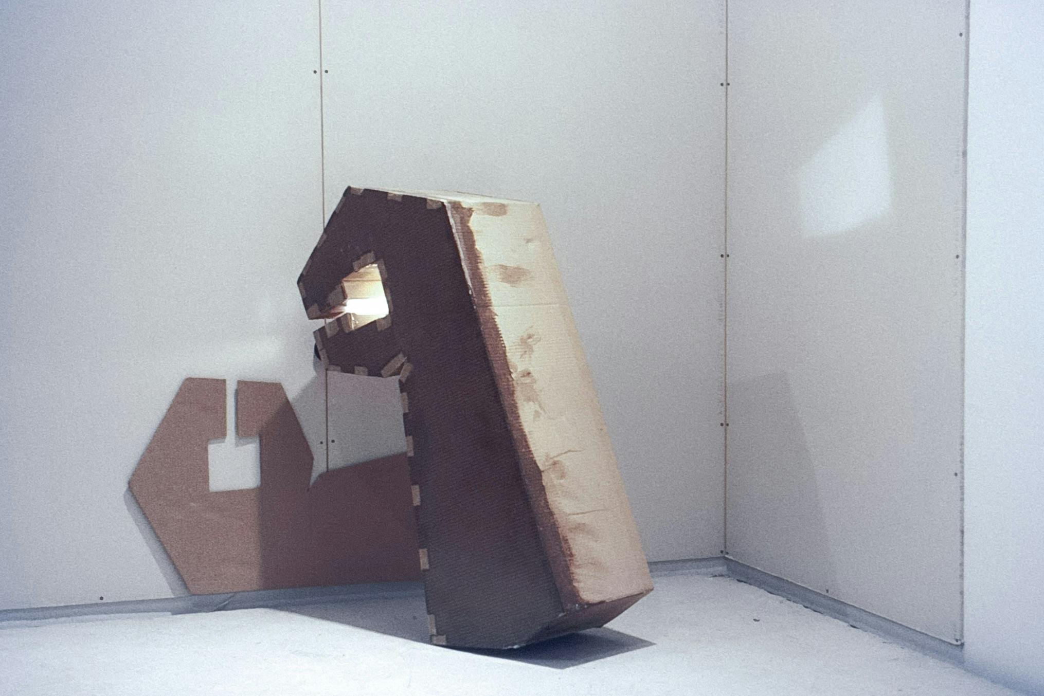 The corner of a gallery with 3 grey panels on the walls. Across 2 panels there is a flat, wrench-shaped cardboard piece. This shape is repeated in 3D, leaning on the wall with a light in its "jaw." 