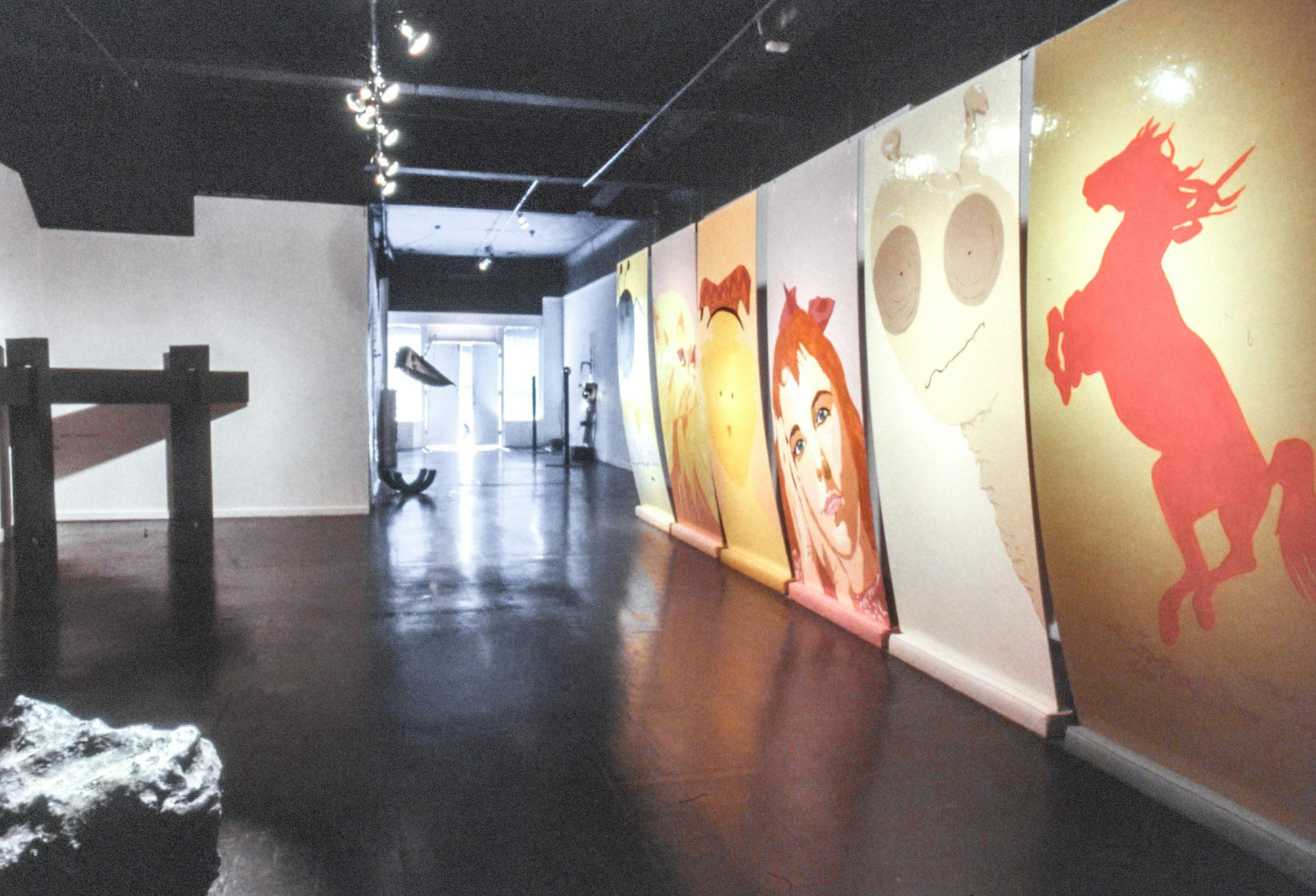 Multiple artworks visible in a gallery. Several colourful wall-to-ceiling paintings on glossy surfaces are visible on one side of the space. The rest of the space has various sculptures all around.