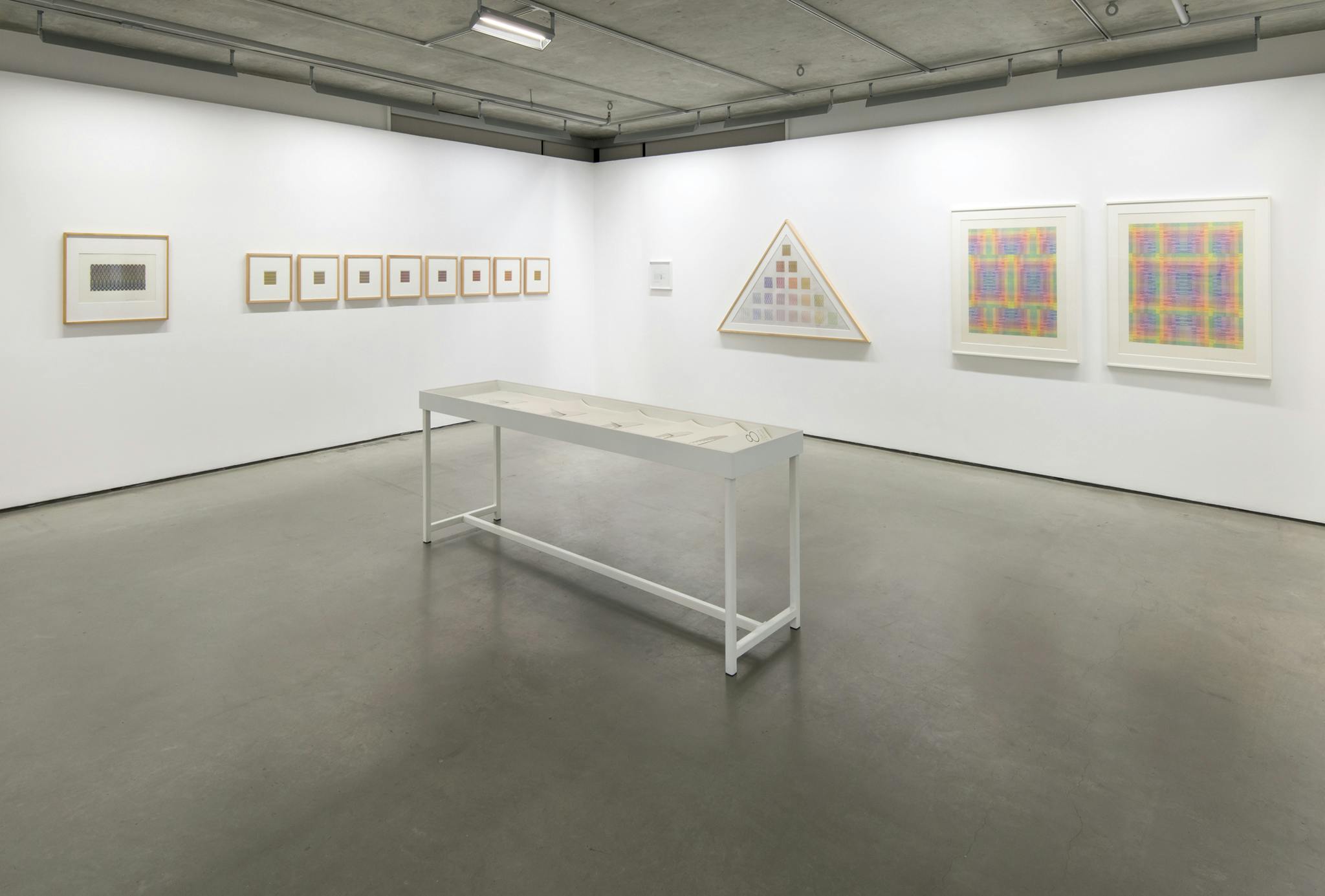 An installation image of artist Channa Horwitz’s exhibition at CAG. Thirteen framed drawings of varying size and shape hang on the wall. A display vitrine sits in the center of the room. 