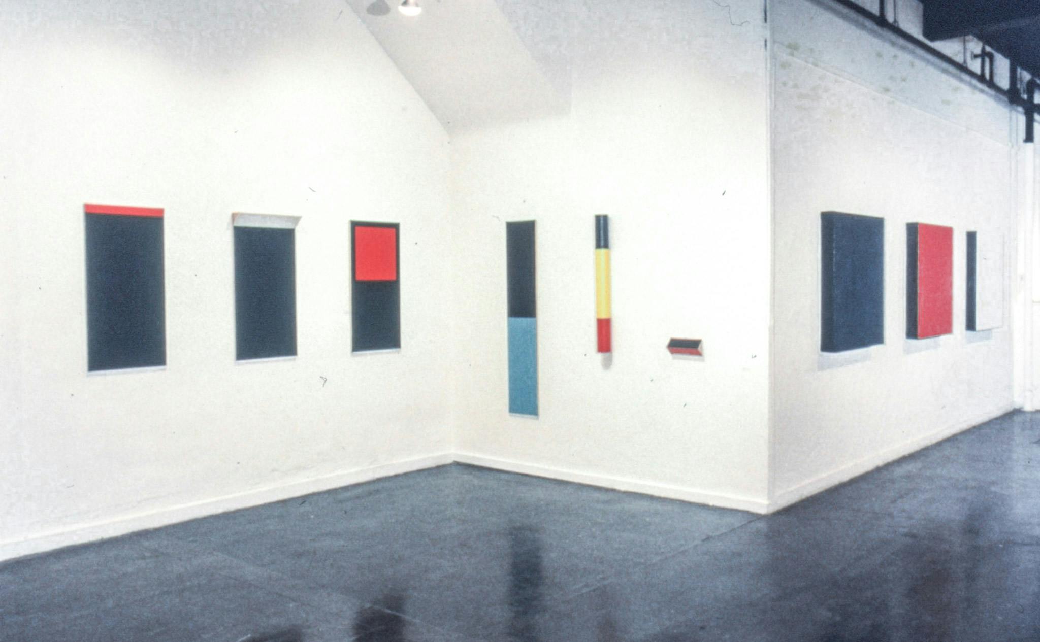 Paintings mounted on three different walls of a gallery. Most are on rectangular canvases, but one painting is a vertical cylinder shape. The paintings are white, blue, red, yellow, and black. 