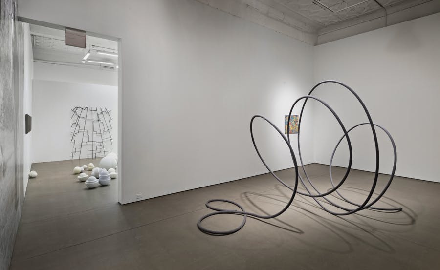 In the front gallery, a coil-shaped black sculpture is installed on the floor in front of small  colourful paintings mounted on the wall. More sculptures are installed in the other gallery