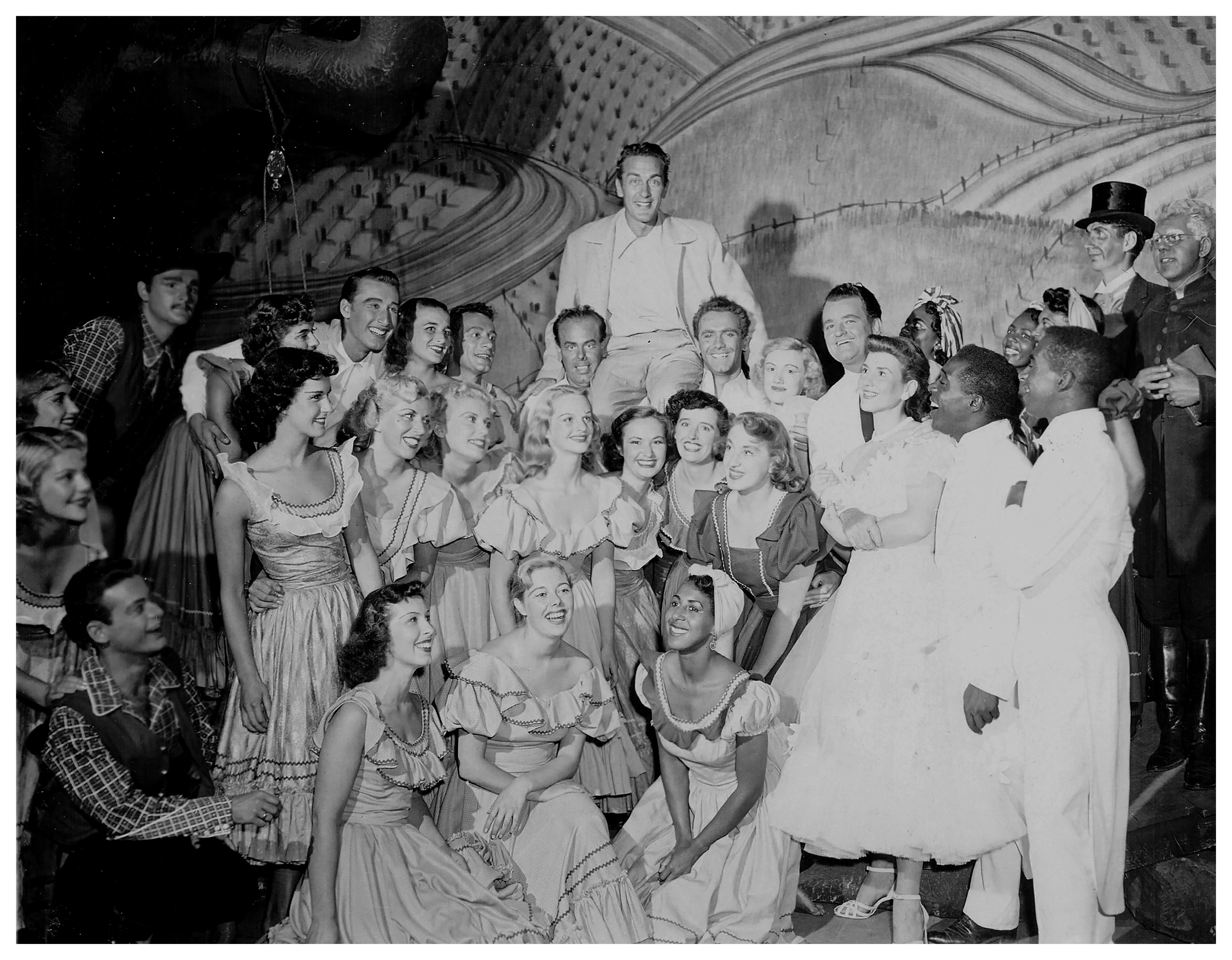 A black and white cast photo from a theatre play with a large group of smiling people standing and crouching in rows. The people appear to be in costume and positioned in front of the stage backdrop. 