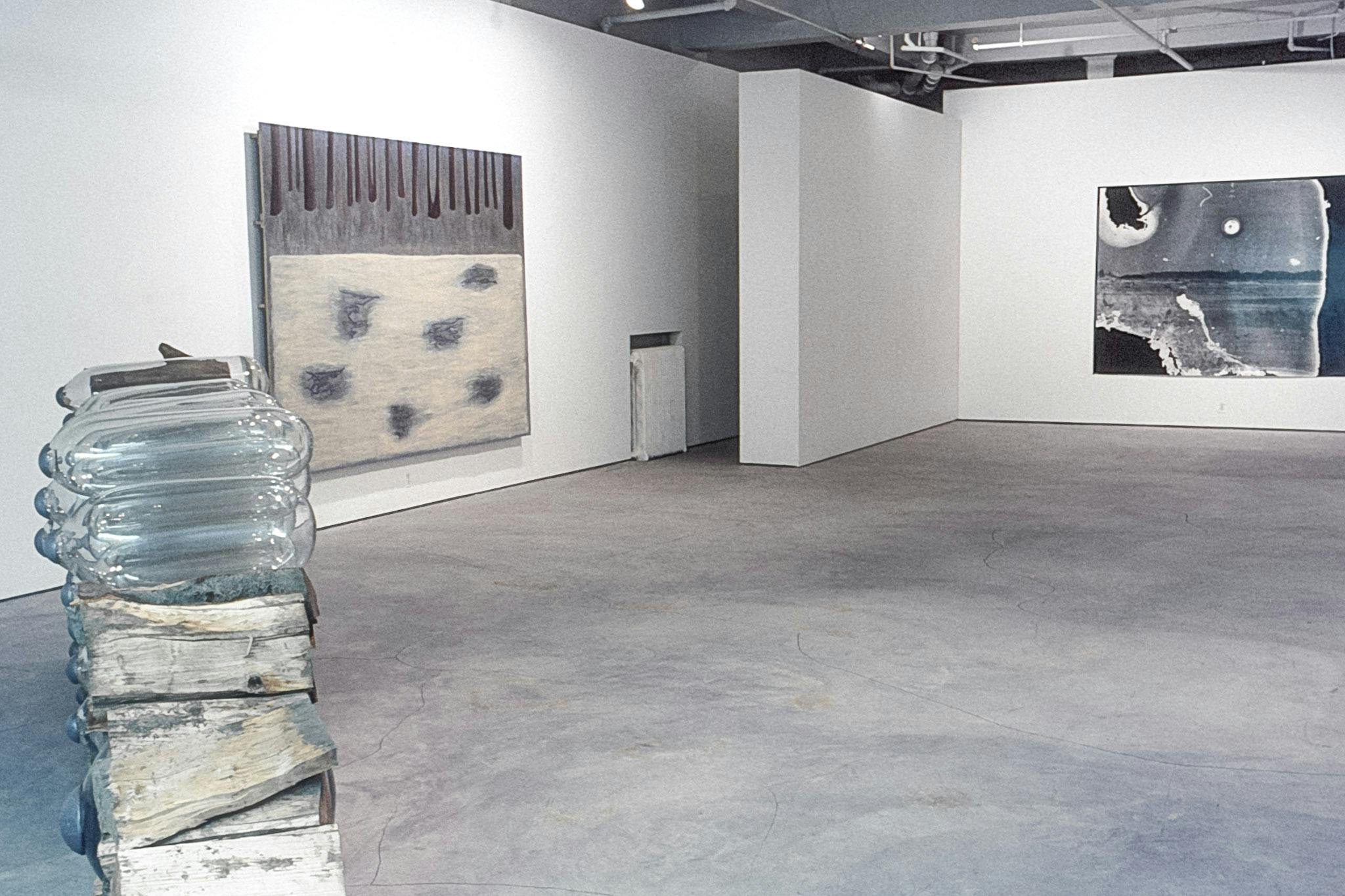 A room in a gallery shows 3 artworks. One is a thin stack of firewood and glass cylinders. One is an abstract yellow, grey and black painting, The other is a large, deteriorated photo of a landscape.