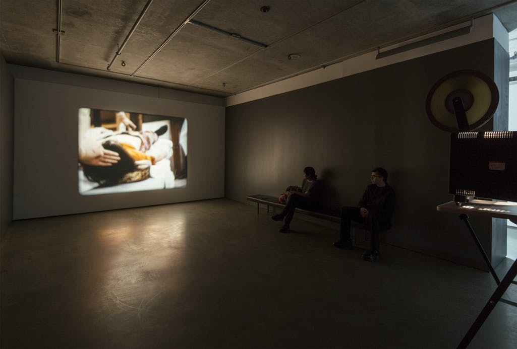 A single-channel video piece is projected on a gallery wall. The video depicts a figure lying on a flat surface. A hand touches the figure’s head. Two visitors sit on a bench to watch this video.  