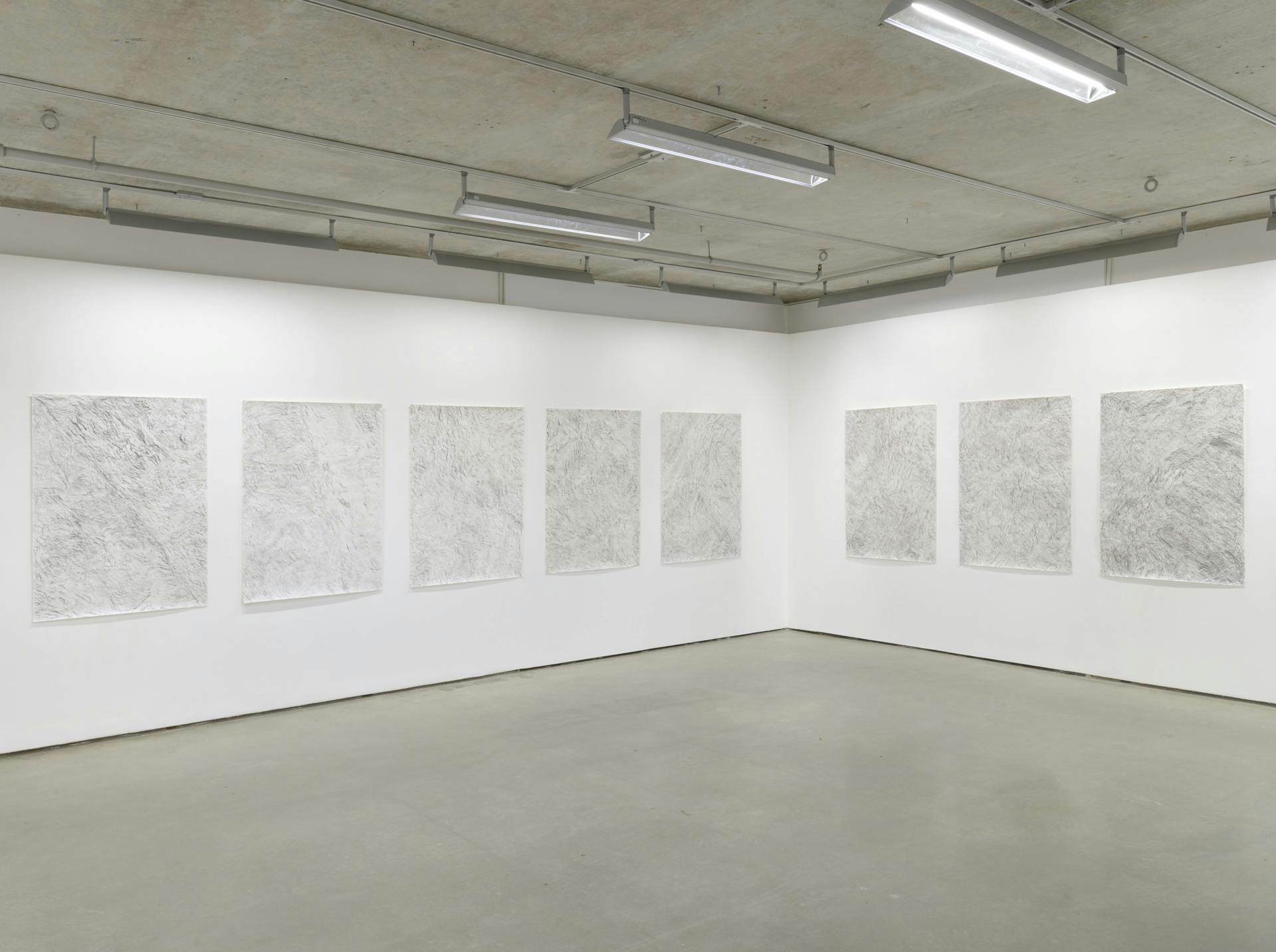 Graphite rubbings on rectangular sheets of paper arranged in a row around a corner in a room with white walls.