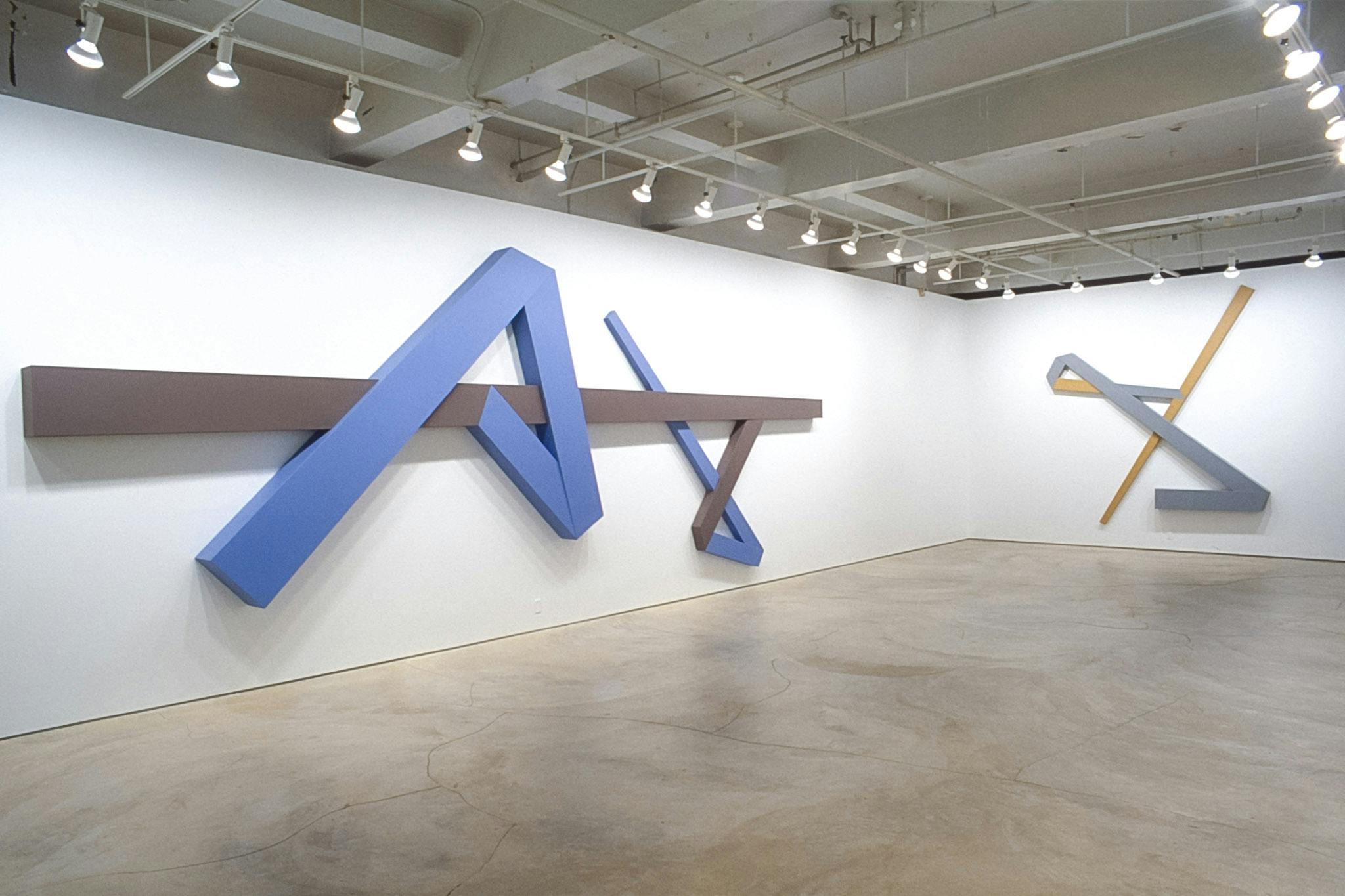 Geometric sculptures by Daniel Congdon mounted on the wall of a gallery space. The visible sculptures are dark-brown with blue, and light-grey with yellow.