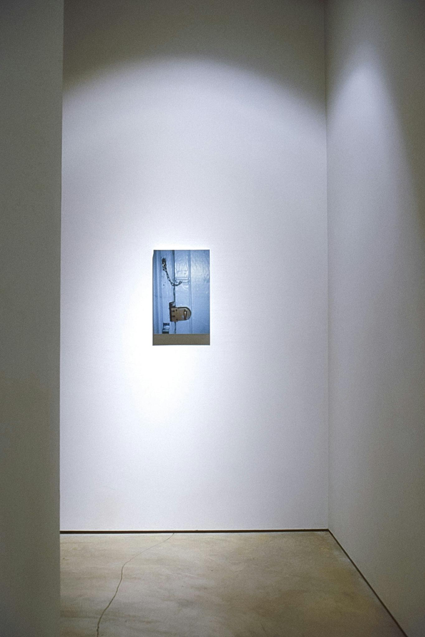 A view of a gallery corner shows a small photograph mounted on the wall. The piece shows a part of a light blue door with a metal lock and a chain lock.