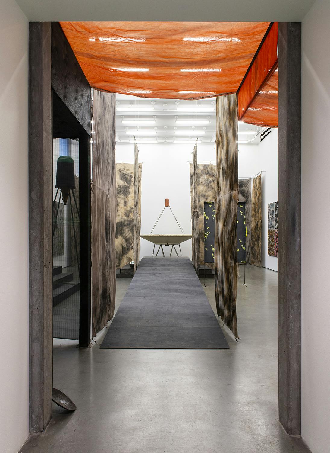 A view of Corbin Union’s exhibition in a gallery space. Long strips of brown and black tie dyed cloth hang throughout the gallery space. A ramp sits in the center of the room and leads to a sculpture.