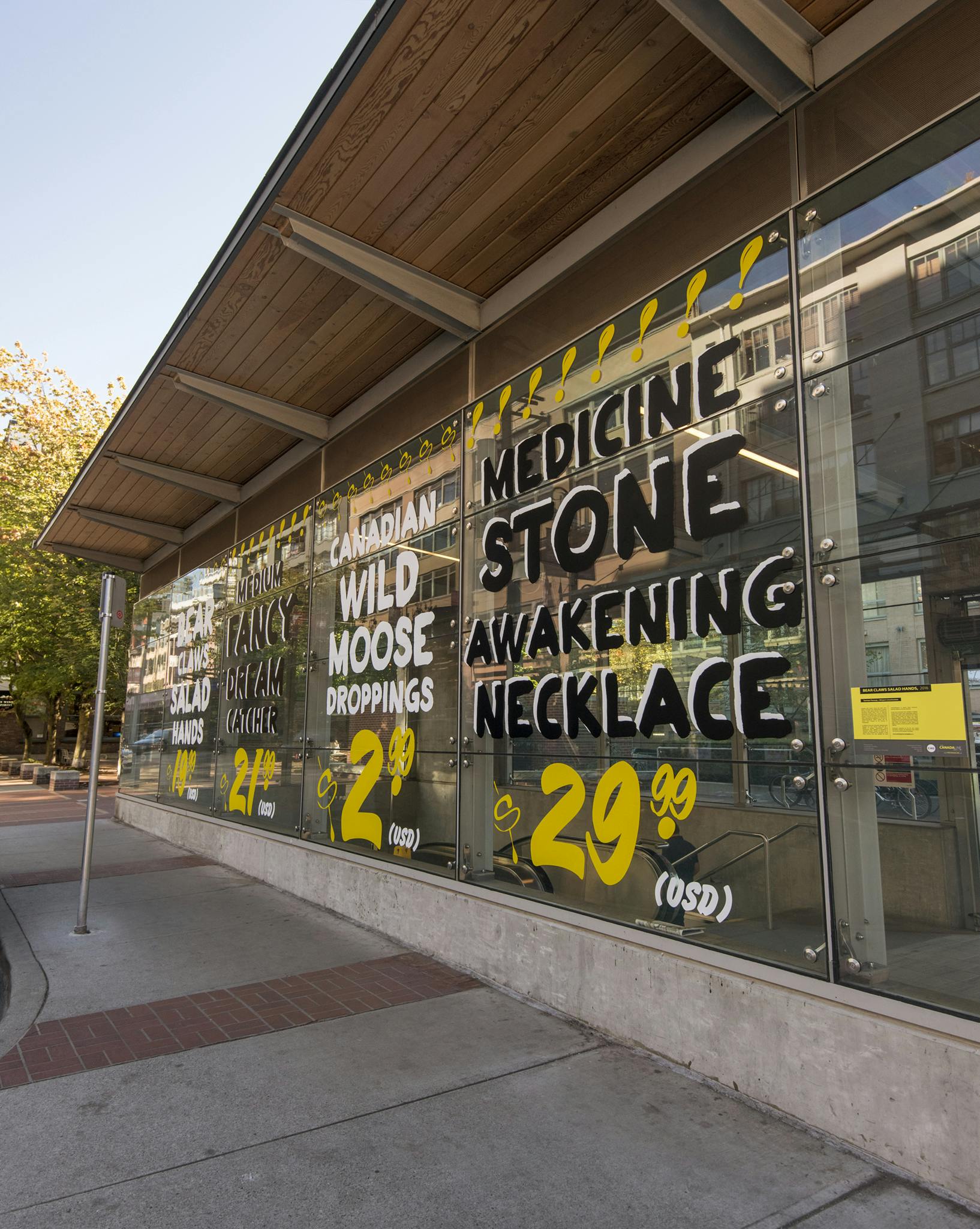 Text-based artworks in vinyl are installed on the glass facade of Yaletown-Roundhouse Station, resembling hand painted signs. One reads: “MEDICINE STONE AWAKENING NECKLACE $29.99 (USD).”