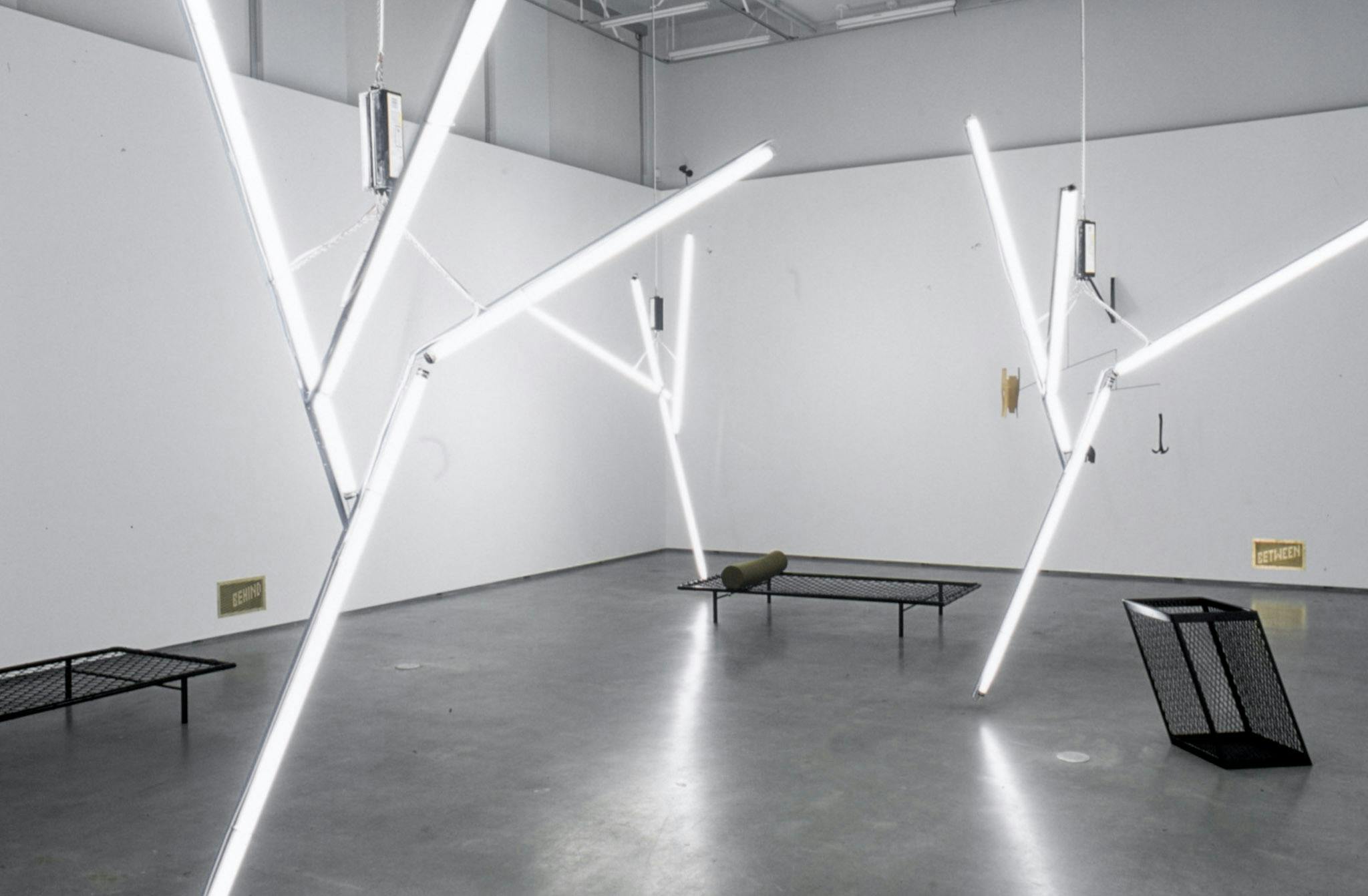 Three large sized sculptures are hanging from the ceiling of a gallery space. They are made of white tube light bulbs that are assembled to look like tree branches. 