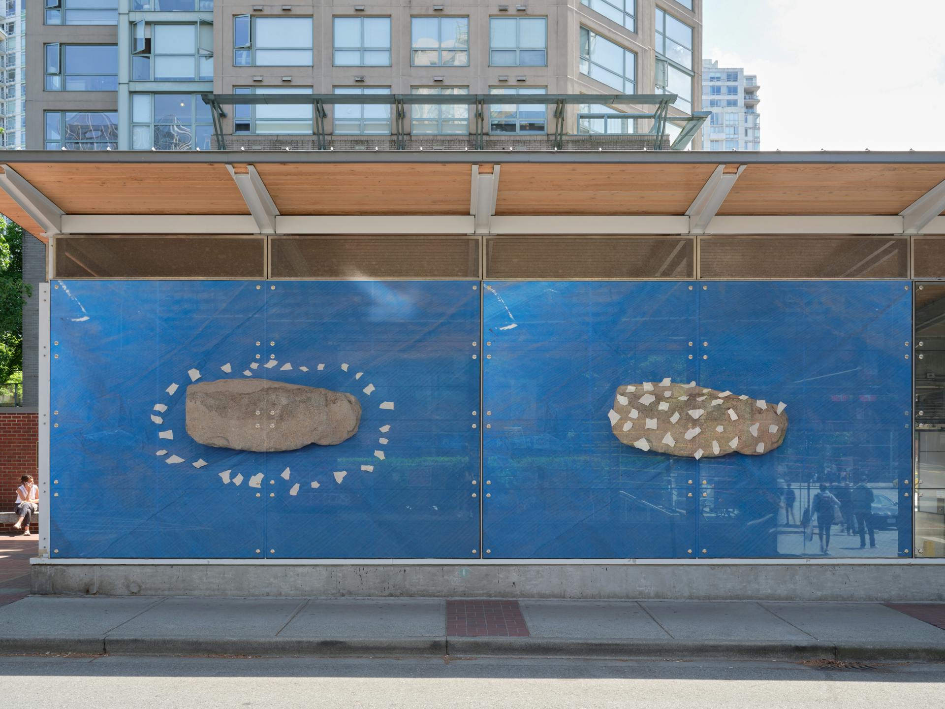 A frontal view of a SkyTrain station with two photos in its windows. The first photo depicts a rock with bits of white tape arranged in an oval around its perimeter. The second depicts the same rock with bits of white tape dotting its surface.