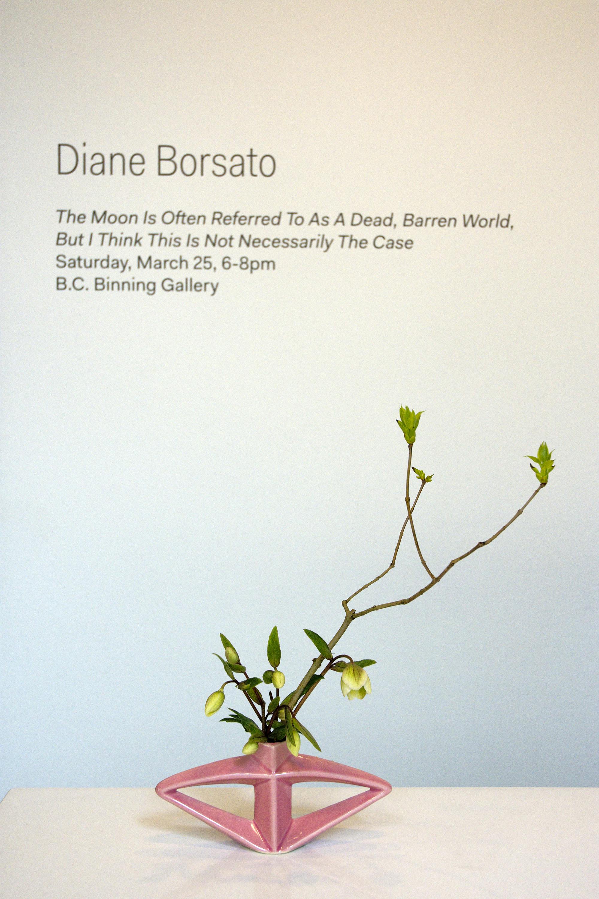 Artist name, exhibition title, date, and location are printed on a wall above a white table. On the table, A triangular-shaped pink vase sits, in which some green flowers and twigs are arranged.